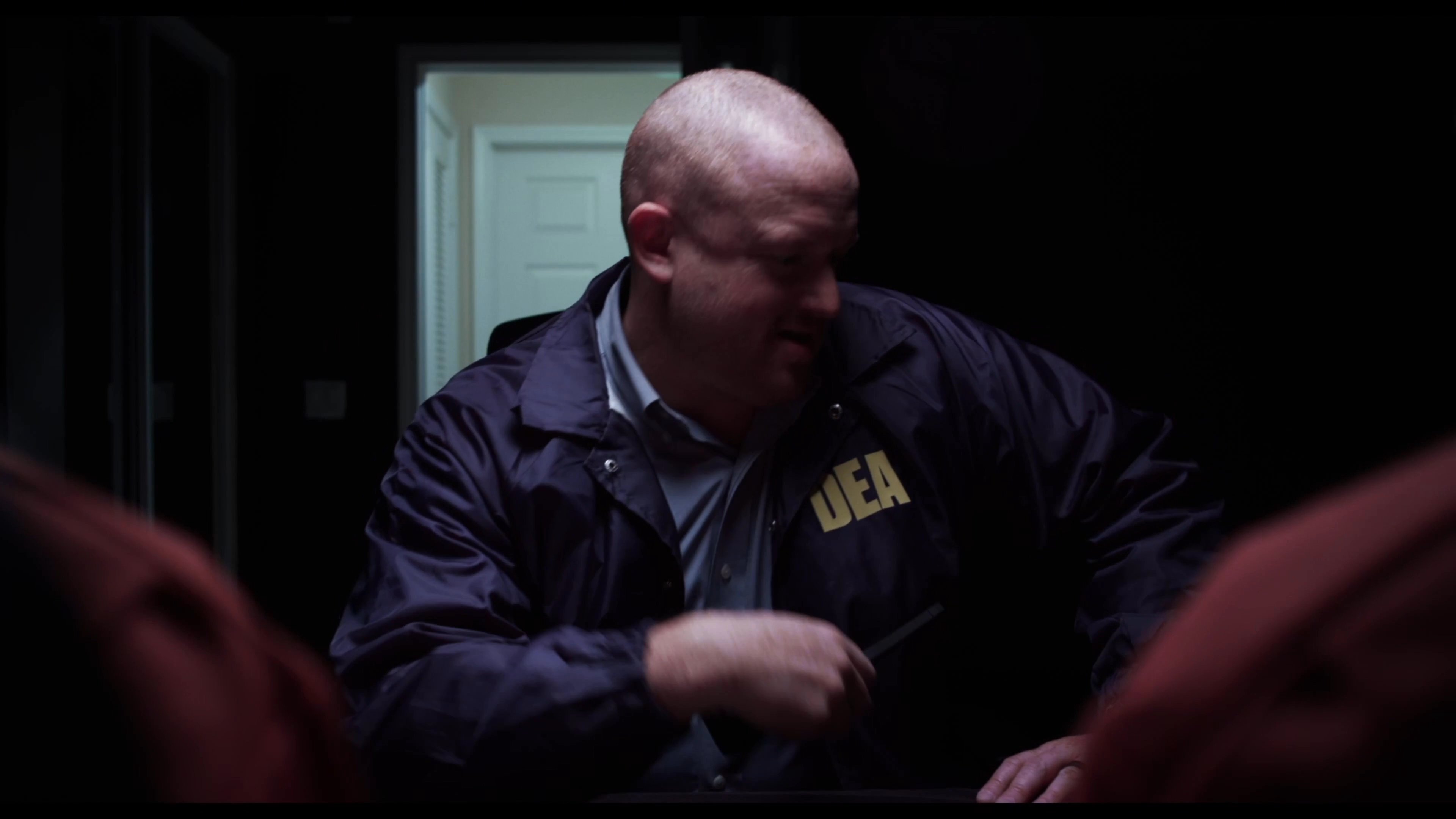 A DEA agent monitors a subject through surveillance equipment during an interrogation process. The officer coordinates activities via two-way radio while observing the suspect's movements.