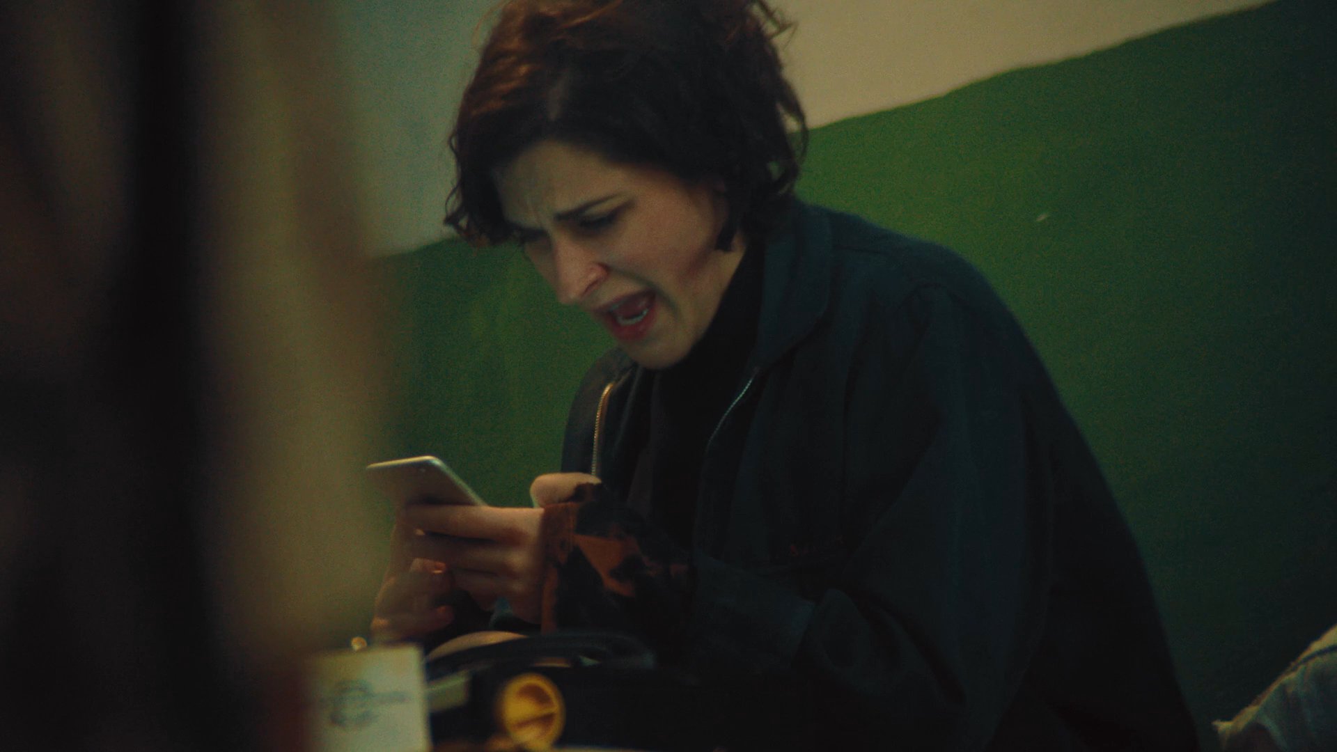 A woman sits in a dimly lit, cluttered room using a smartphone before transitioning to a private moment in bed. The scene captures an intimate sequence of individual activity and relaxation.