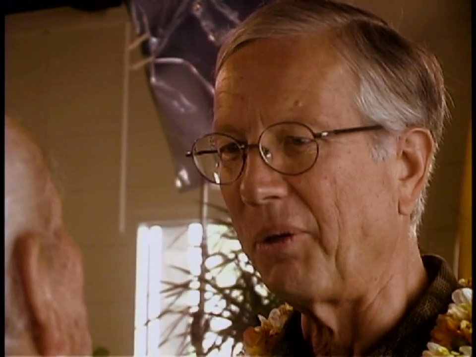 An older man wearing a floral lei engages in a friendly, animated conversation with an elderly gentleman and a woman during an indoor social event.