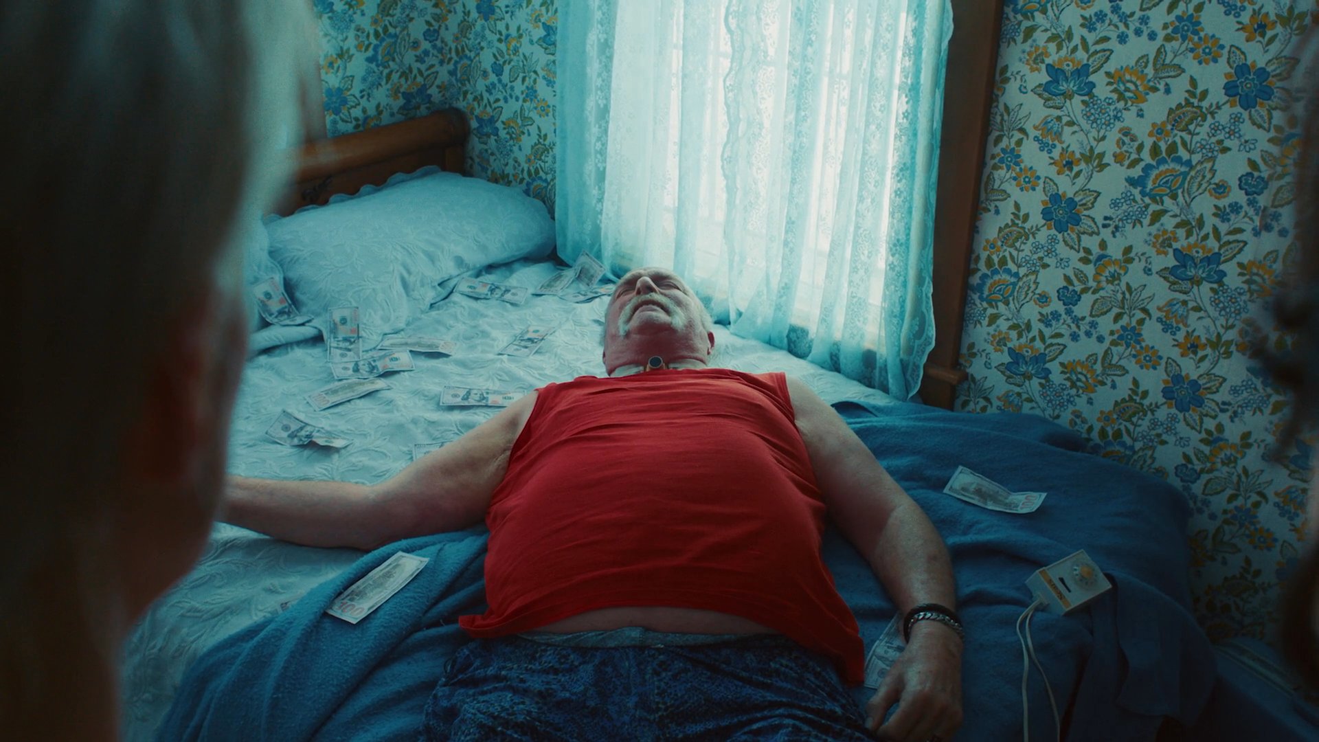 Two individuals stand in a cluttered bedroom while observing an older man lying on the bed. The scene is marked by a palpable sense of tension and unresolved conflict.