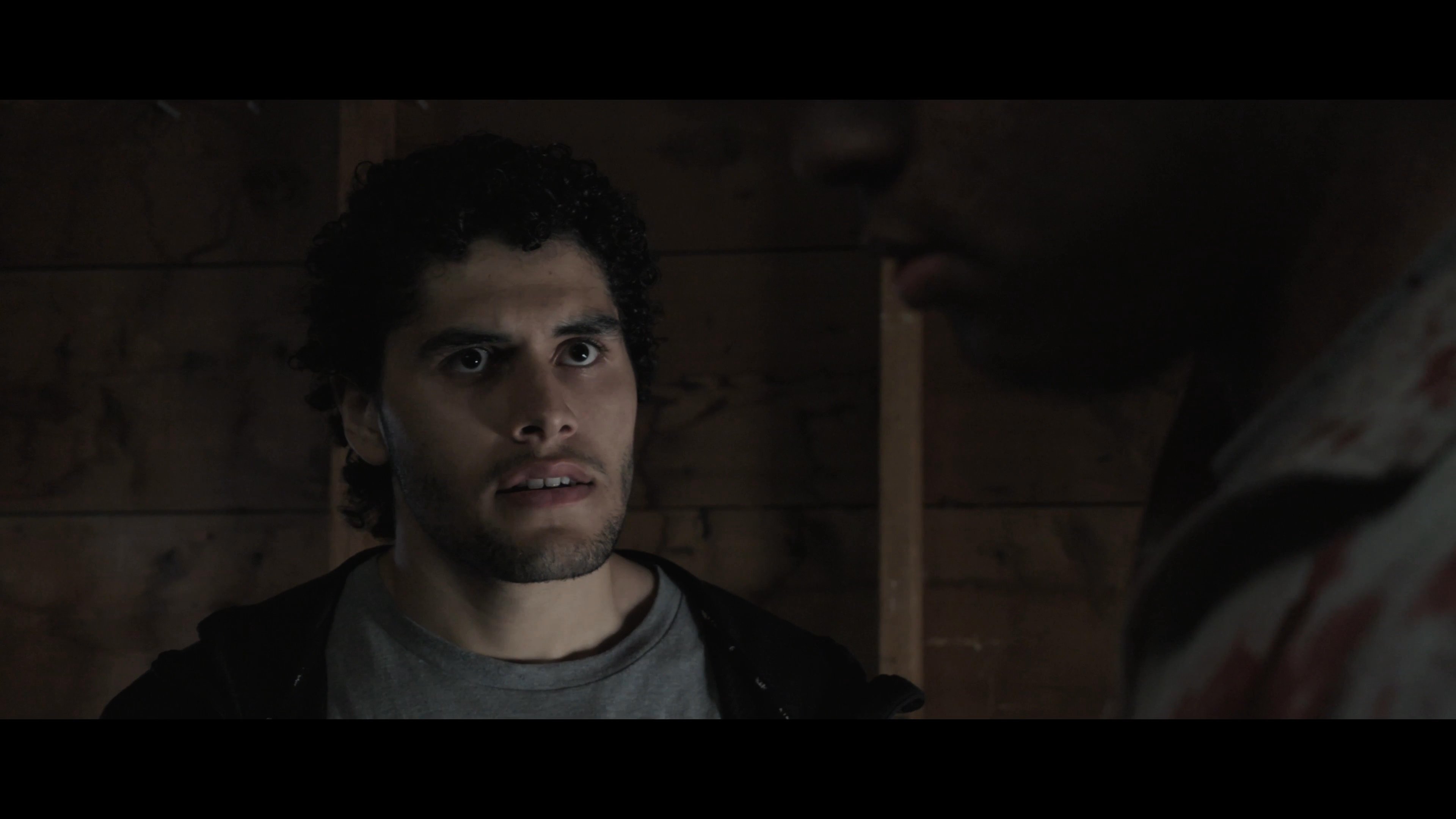 Two individuals engage in a tense, emotionally charged exchange within a dimly lit, rustic structure. The dialogue appears critical, with both parties demonstrating significant distress and focus during the interaction.