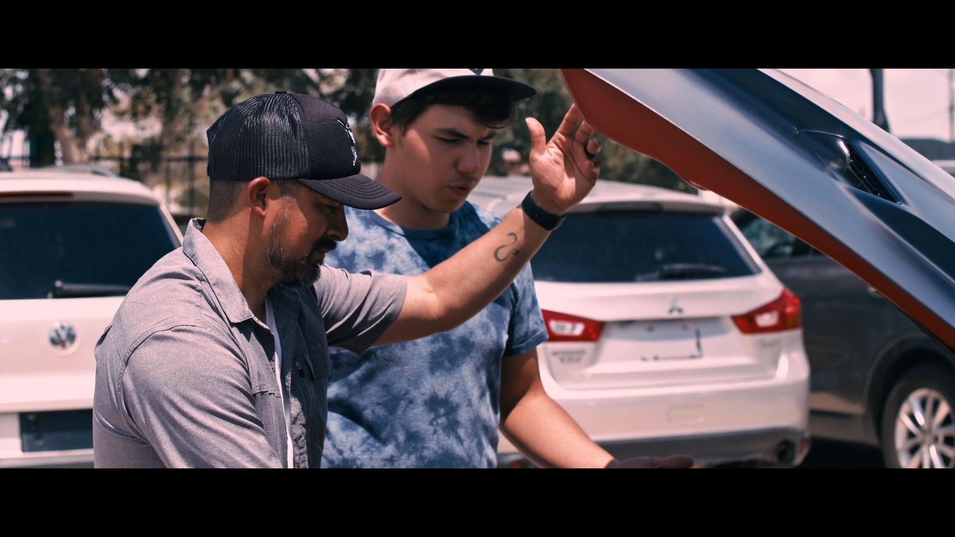 Two individuals inspect the engine compartment of a red Dodge Challenger in an outdoor parking area. They discuss mechanical aspects of the vehicle while examining the engine bay.