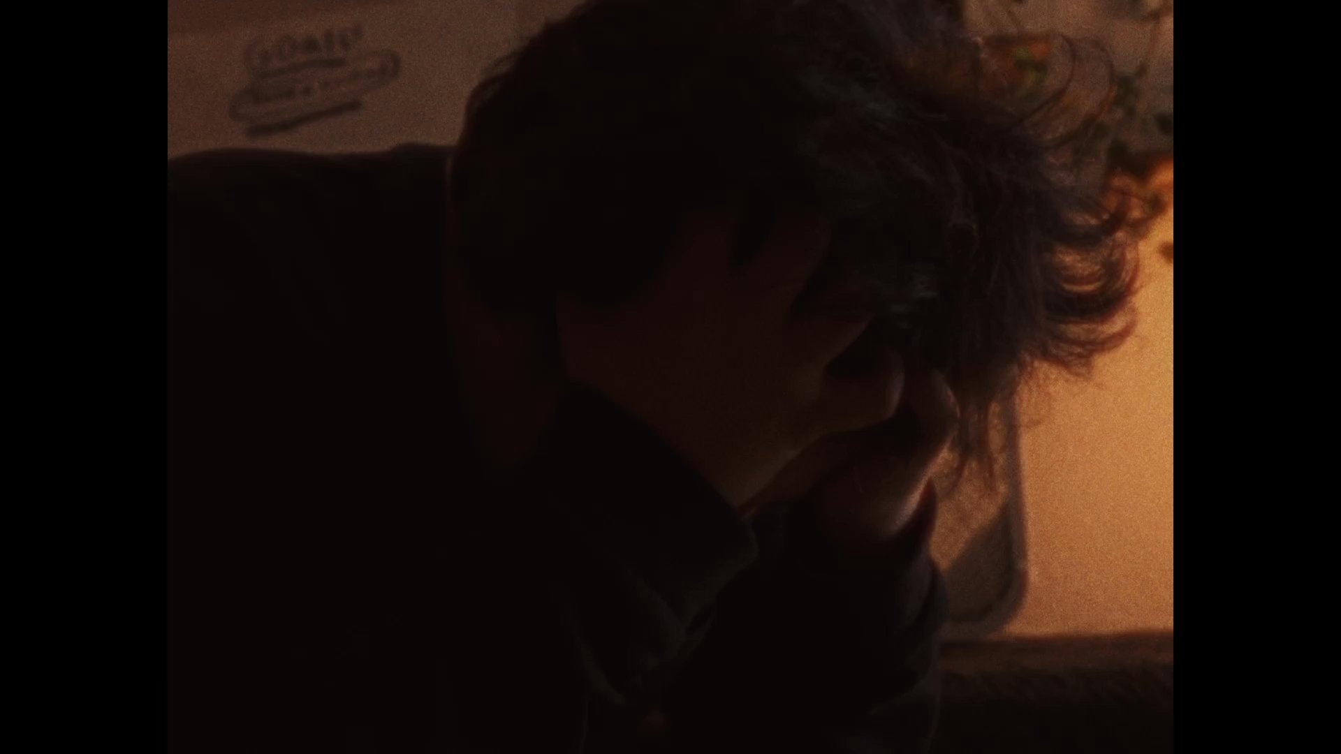 A young individual sits in a dim, amber-lit room, clasping their hands over their face while exhibiting signs of deep contemplation or emotional distress.