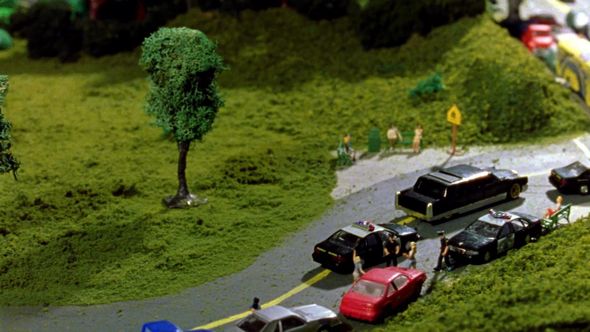 Pedestrians walk through a park as a traffic light transitions from green to red, overlooking an intricate miniature intersection filled with model vehicles. The scene shifts to a contemplative close-up of a young man, contrasting the bustling scale of the model city with his quiet introspection.