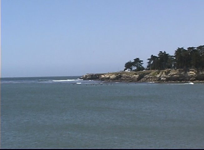 A man provides commentary on a surfing session as the camera pans across a coastal landscape, revealing surfers waiting for waves in the ocean.