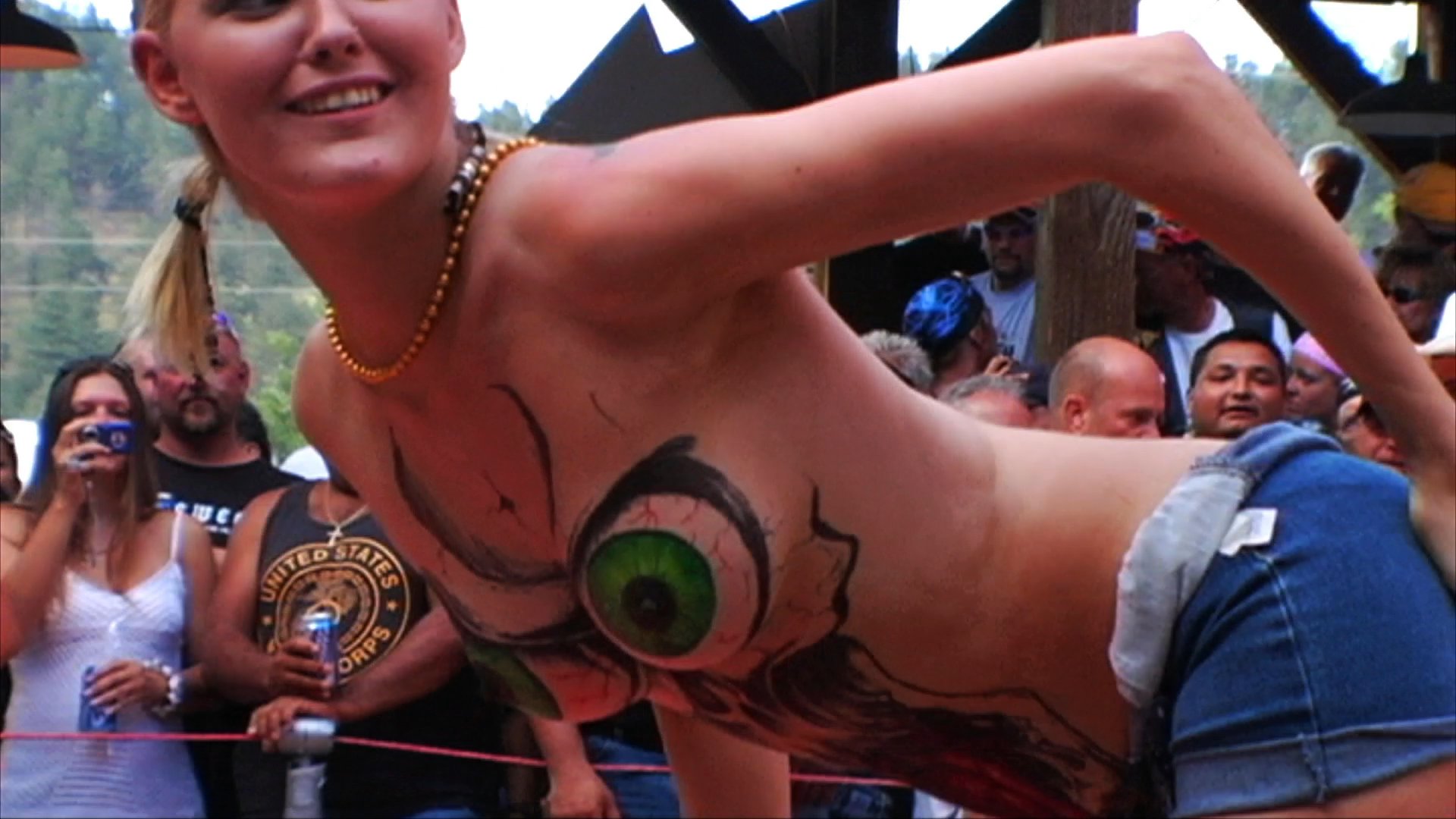Two fighters engage in a boxing exhibition before a large crowd at an outdoor rally, followed by a display of body-painted models and a smoke-filled motorcycle demonstration.