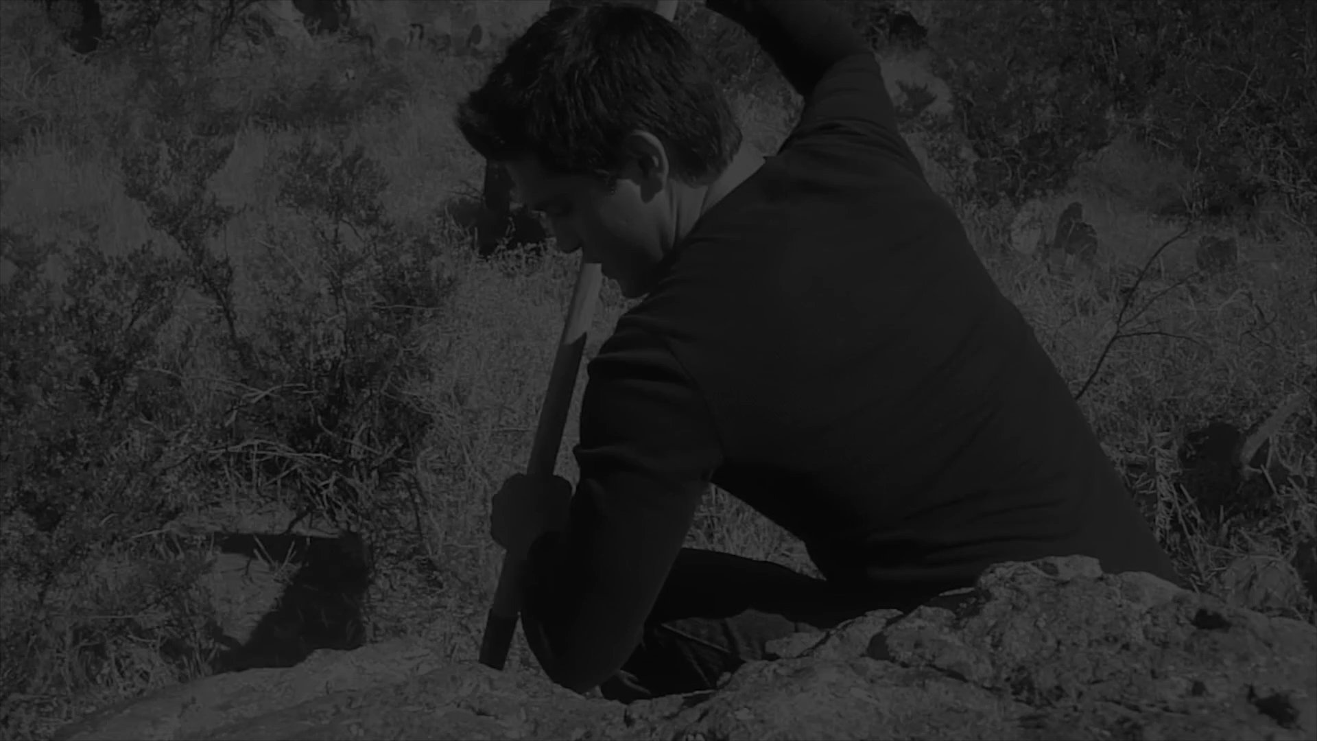 A researcher conducts a manual excavation in a rocky desert landscape, eventually uncovering and retrieving a small wooden box from the earth.