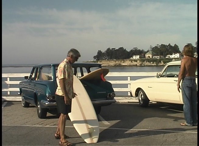 Surfers prepare for an outing, transitioning from vintage car loading to paddling out near a sailboat on the water. The narrative captures the classic aesthetic of coastal surf culture.
