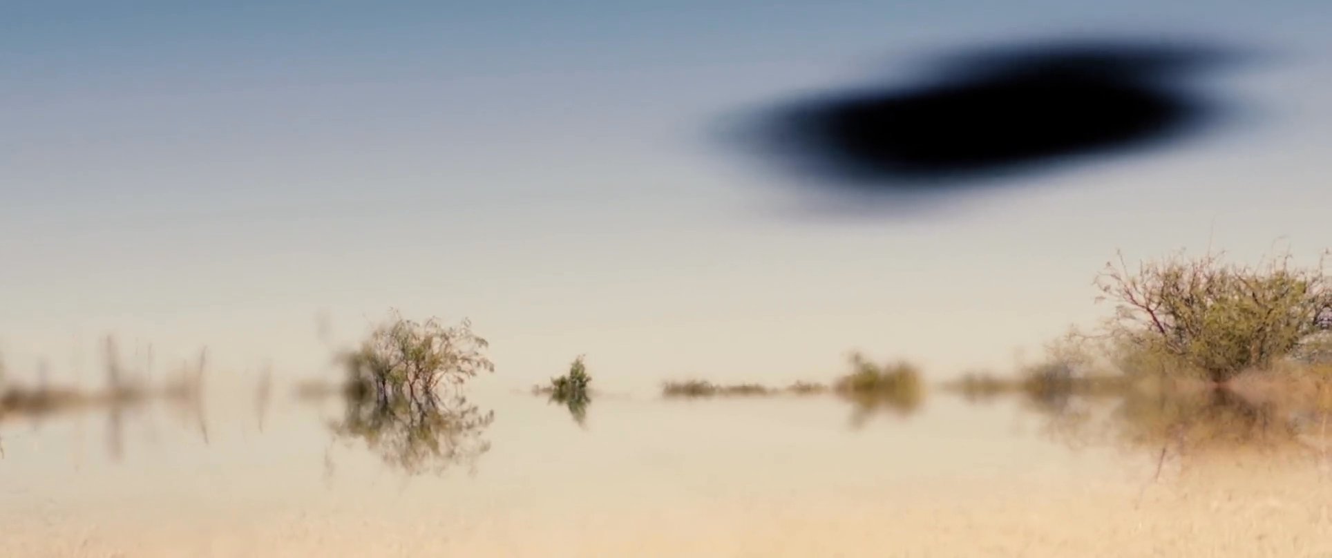 A man stands in an arid, reflective desert landscape, observing a mysterious dark object hovering in the sky. He removes his sunglasses, looking around in confusion as a silhouette appears in the distance.