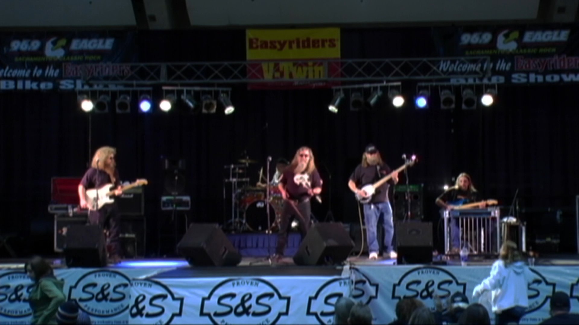 A musical ensemble performs live on stage during a rally event in Leggett, followed by a sequence of motorcyclists riding along a winding forest road.