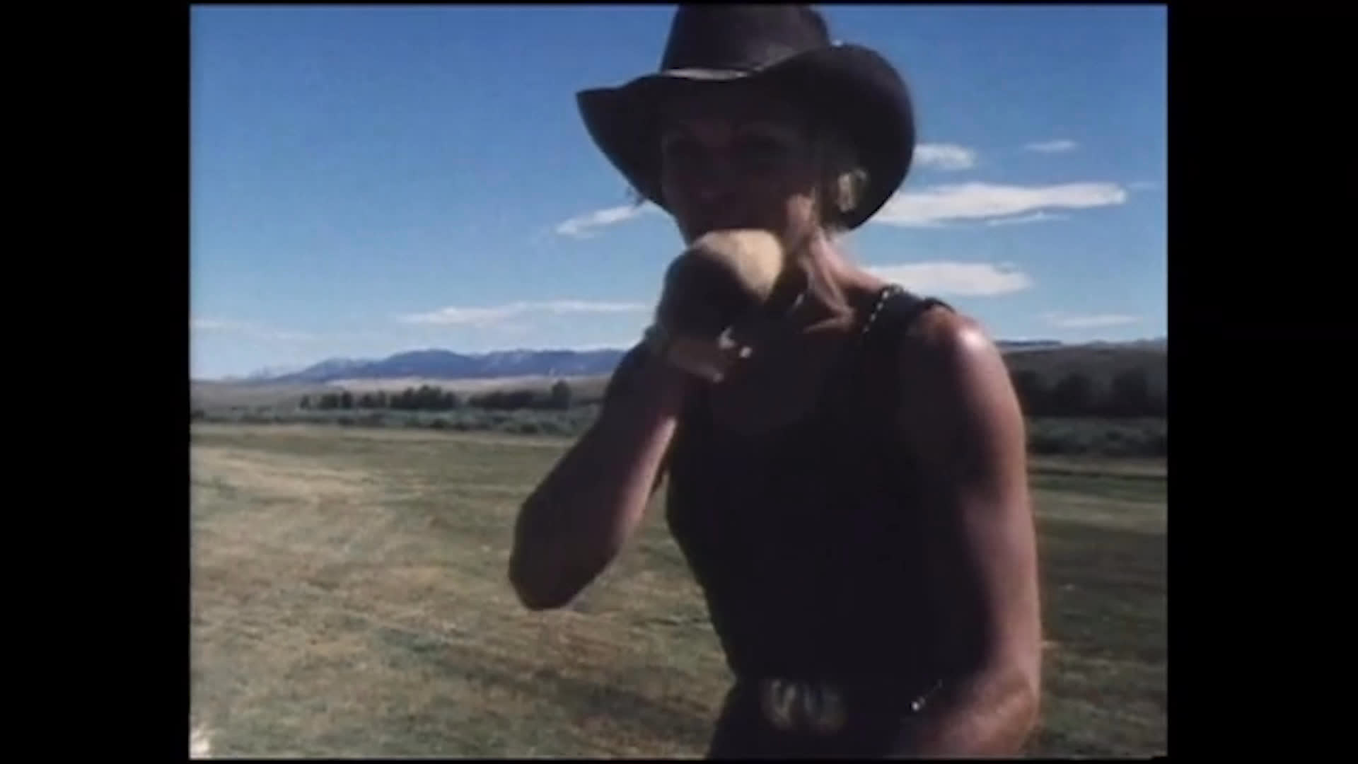A person in a cowboy hat speaks and gestures in a broad, sunlit field with mountains in the distance. They describe workers struggling with heavy workloads and hope a sick worker will return after recovering from the flu.