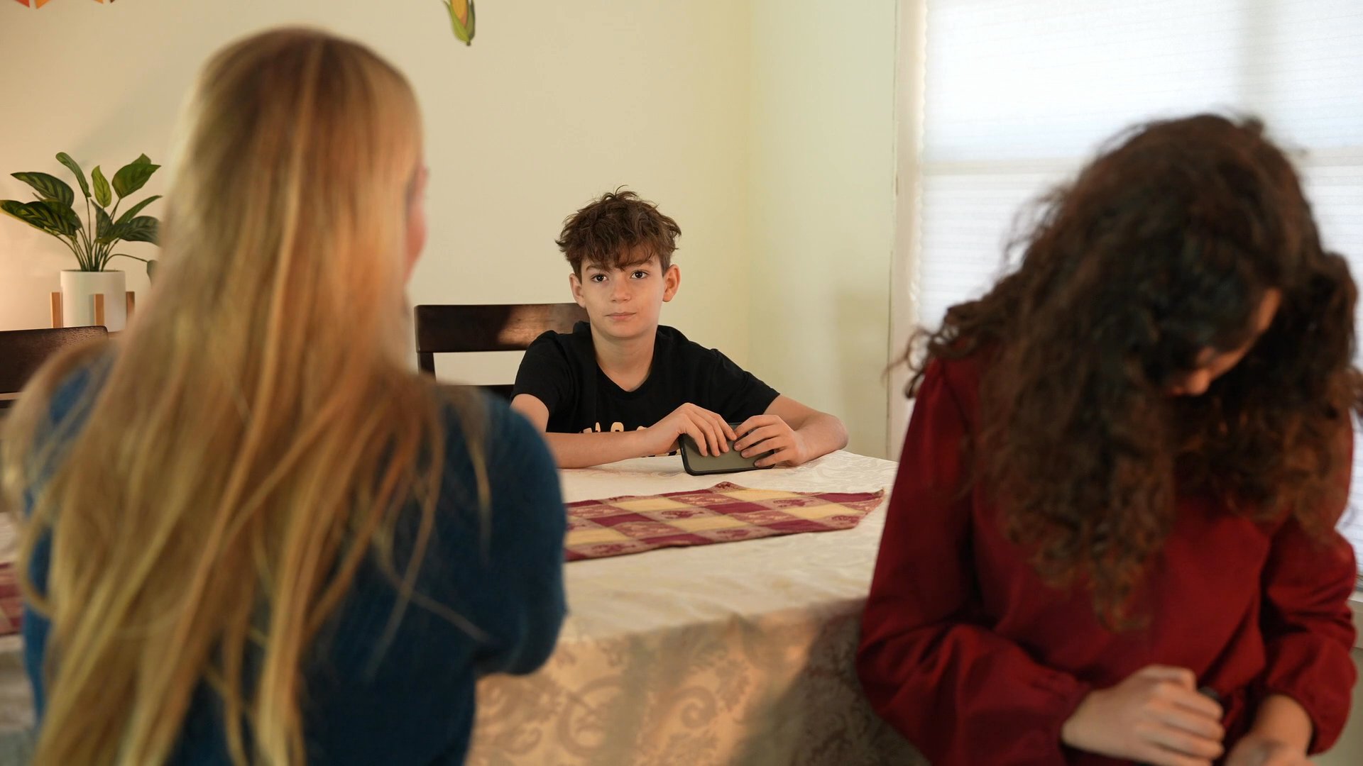 Two women seated at a dining table engage in a focused discussion using hand gestures while a young boy observes in the background. The interaction transitions to an interior kitchen space where a man enters the scene.