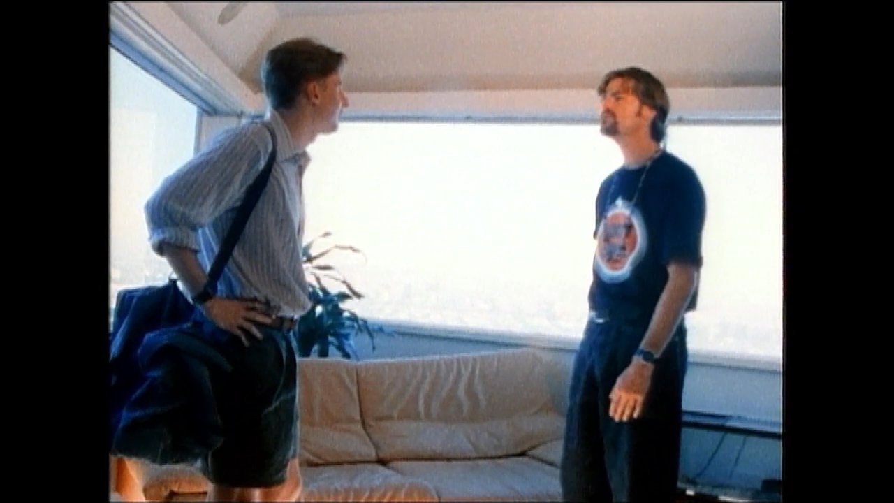 Two men engage in conversation while standing in a brightly lit living room before one of them sits down on the sofa to continue the discussion. The informal atmosphere suggests a friendly social exchange between acquaintances.