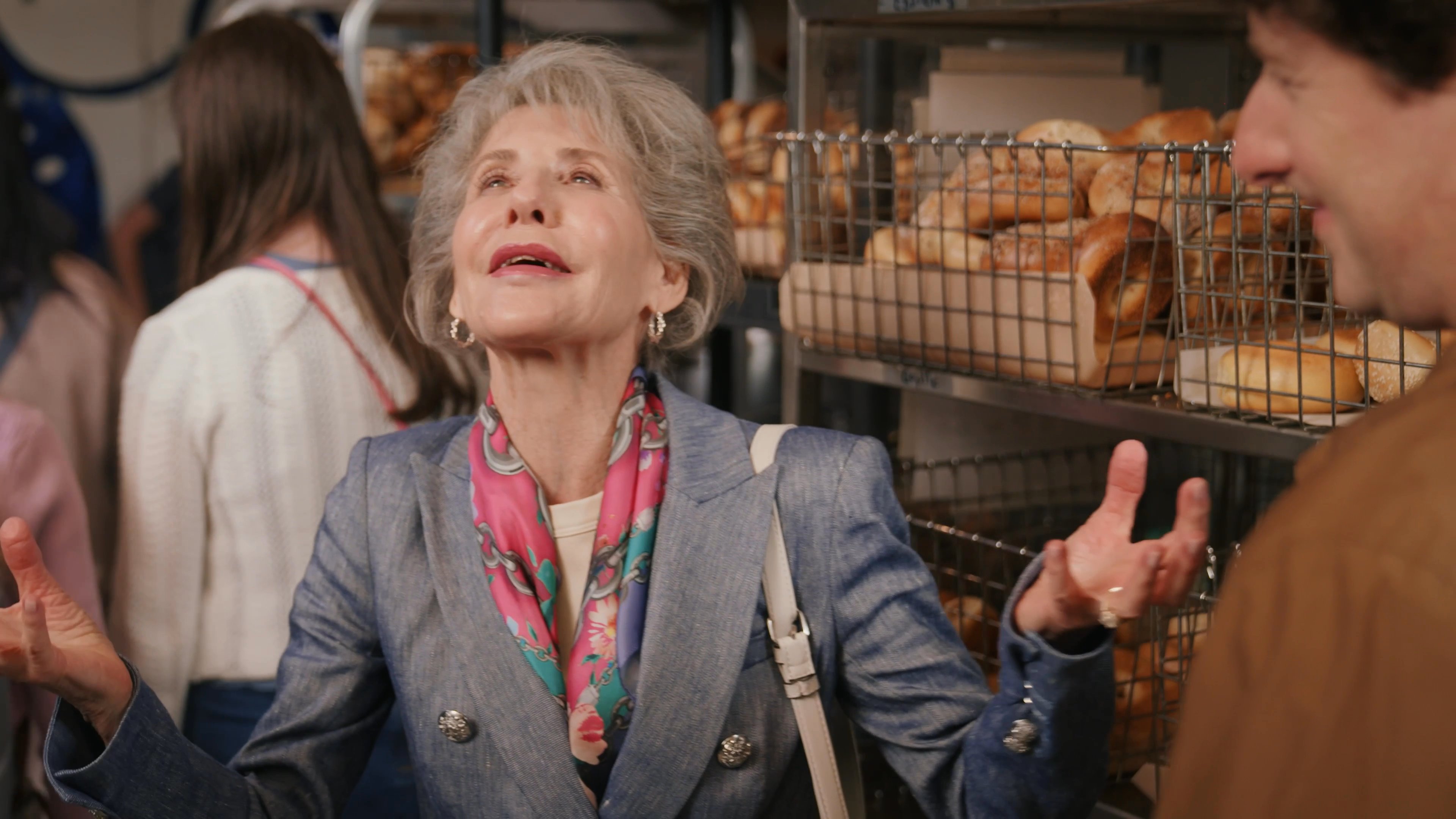 An older woman engages in a lively conversation with a younger man while standing in front of a rack of bagels in a bakery. The two maintain a friendly dialogue in the busy retail environment.