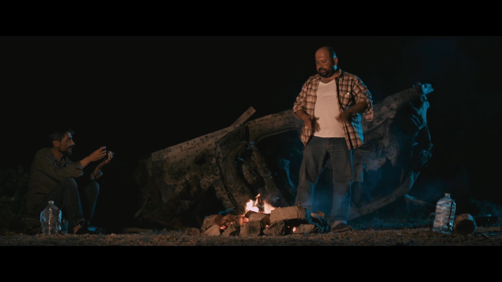 Two men converse beside a campfire in a desolate landscape at night, with one handing a small object to the other as a memento for his child.