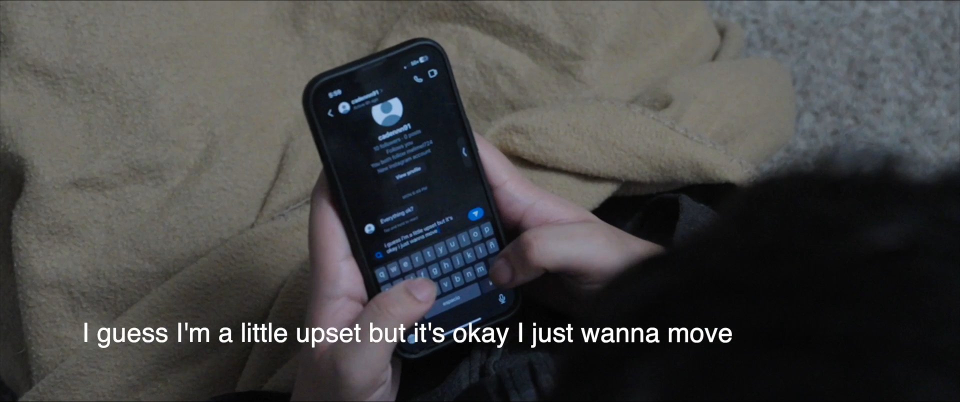 A person types a personal message on a smartphone while sitting in a dim bedroom setting. The individual engages with a social media or messaging interface to communicate their current emotional state.