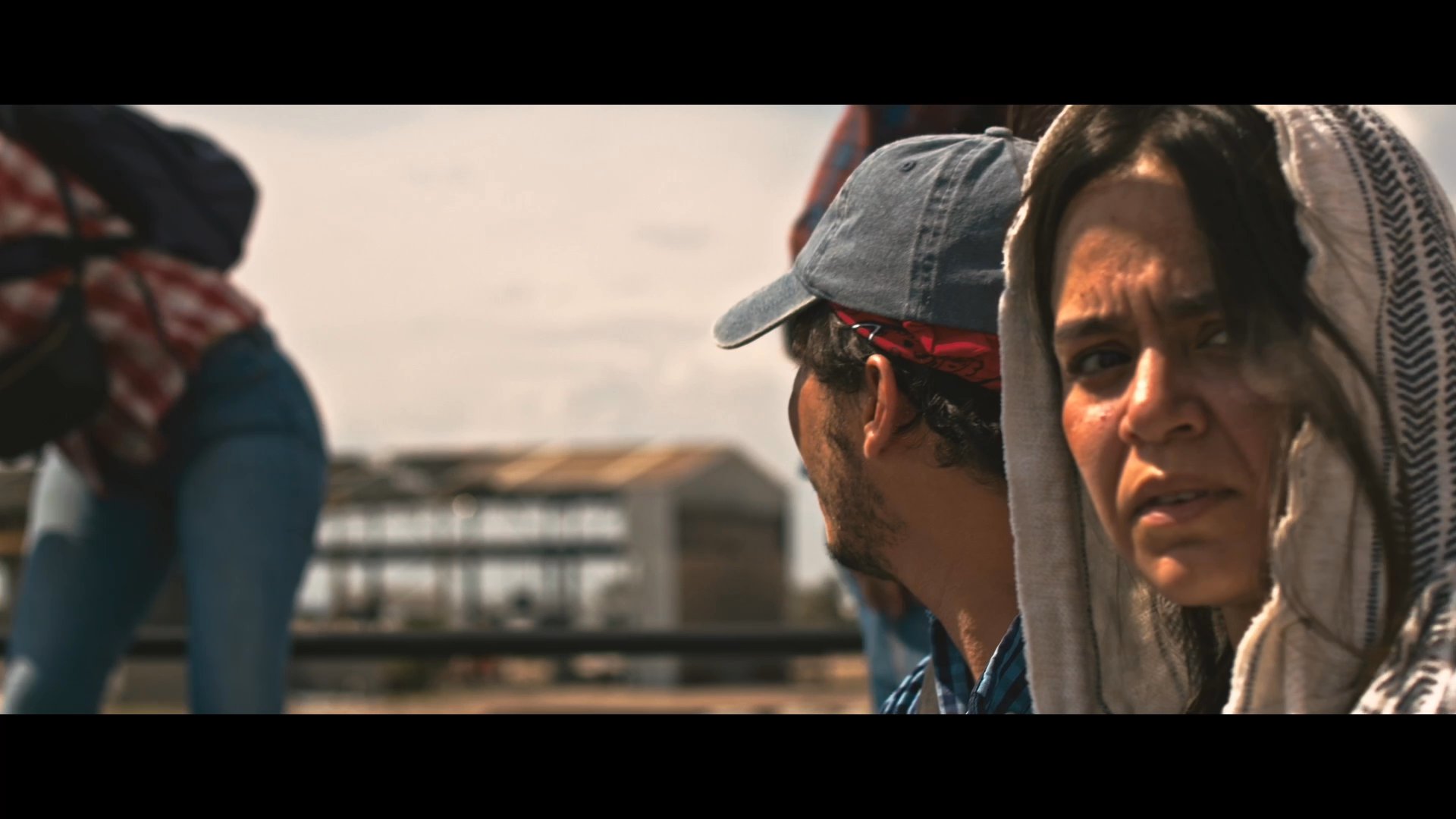 Two travelers exchange conversation while riding atop a freight train through an industrial landscape. The scene contrasts the gritty reality of their journey with a brightly painted mural backdrop.