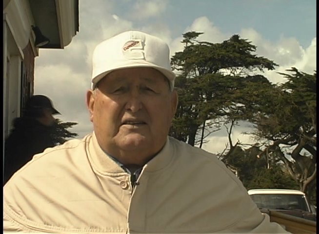 An elderly man shares personal recollections of a coastal location while archival black-and-white footage of a beach pier plays alongside his narration.