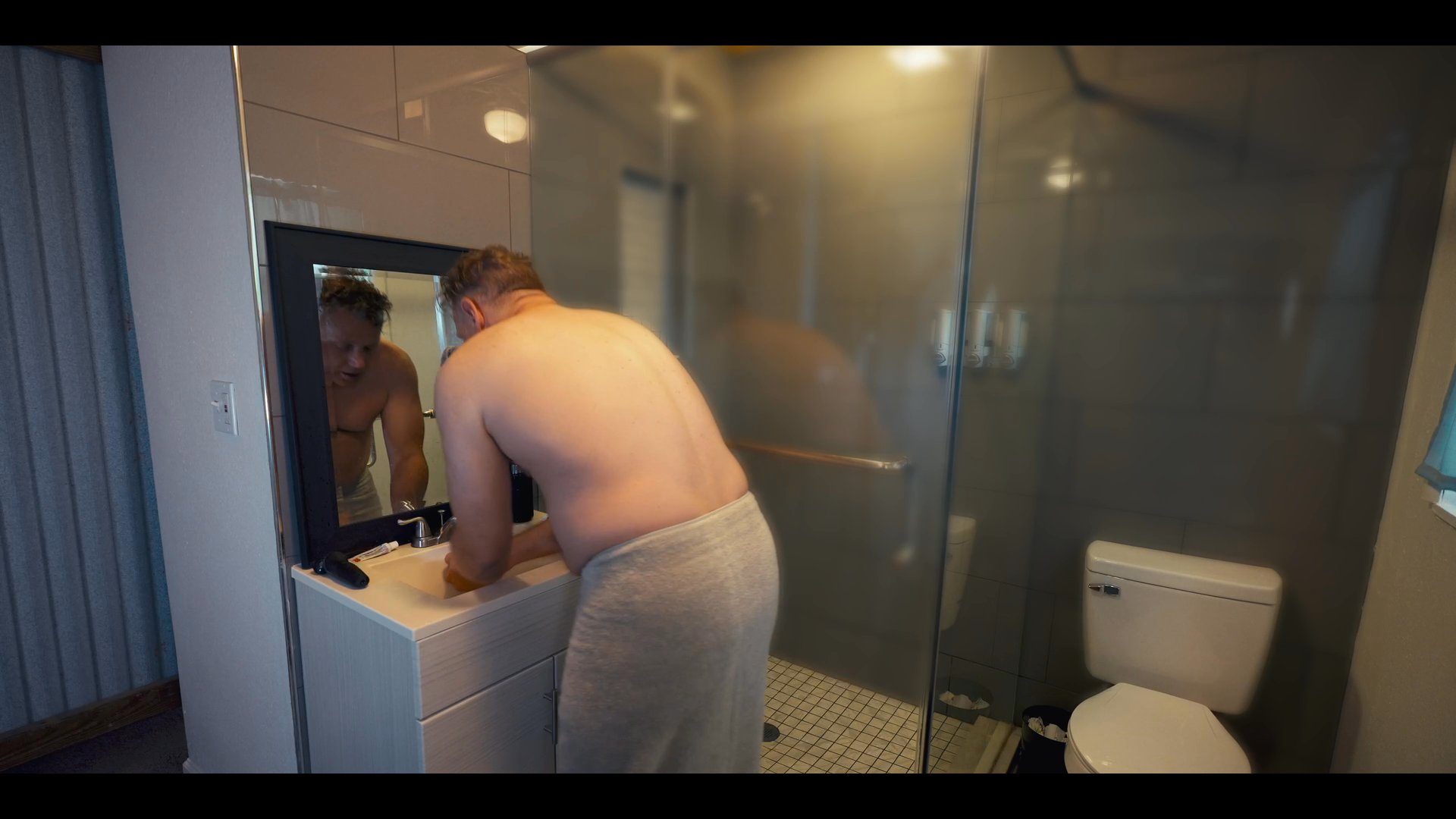 An aerial view of a suburban neighborhood transitions to a man performing his morning grooming routine in a bathroom. He completes his task and walks out into the bedroom area.