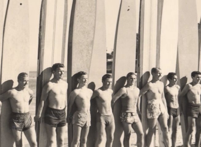 Harry Mayo provides historical context while archival imagery shows a large group of young men posing with vintage surfboards on a beach.