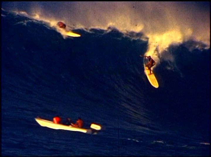 An experienced surfer discusses technical aspects of big wave riding while archival footage demonstrates athletes navigating massive swells and interacting with support watercraft.