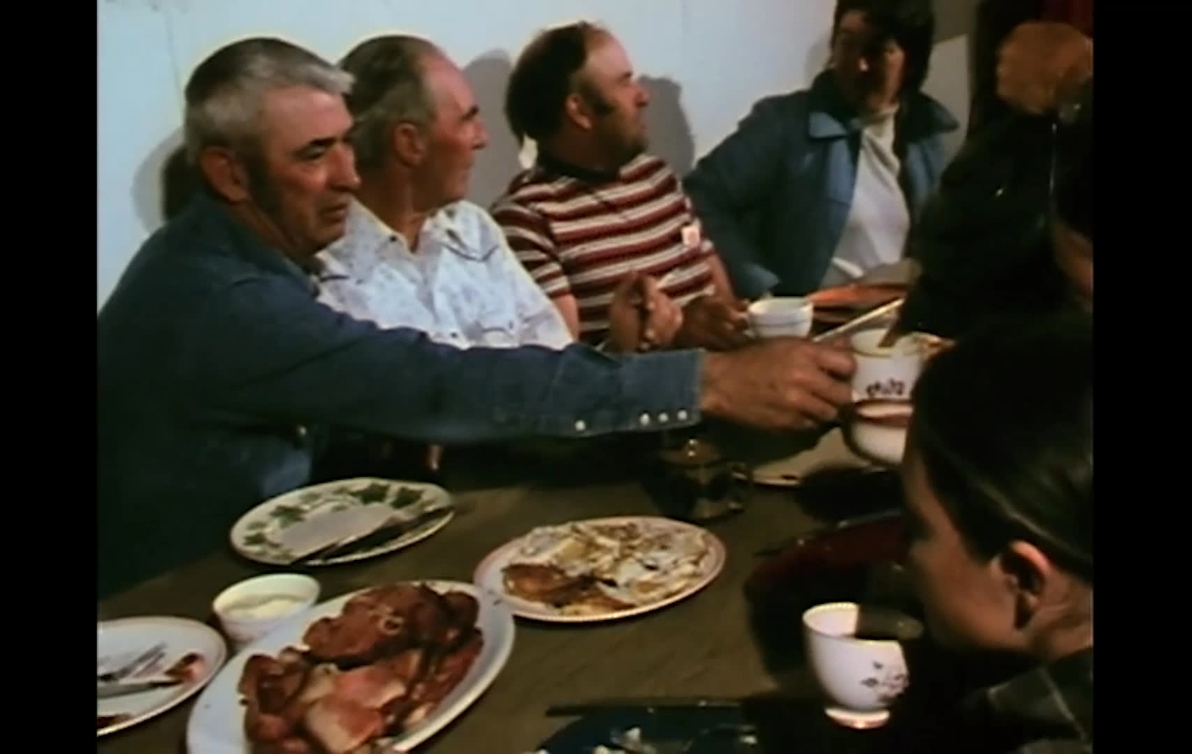 A large pot of coffee boils on a stove before being poured for a group gathered around a table of food. The men eat and visit together, describing the work as a chance to reconnect with friends after the summer.