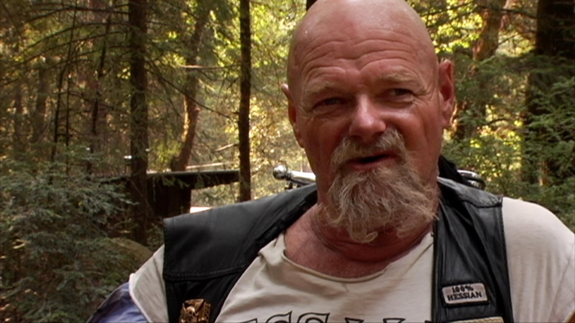 A man with a goatee wearing a leather vest speaks directly to the camera while situated in a wooded environment. He engages in a thoughtful conversation, maintaining steady eye contact throughout the discussion.