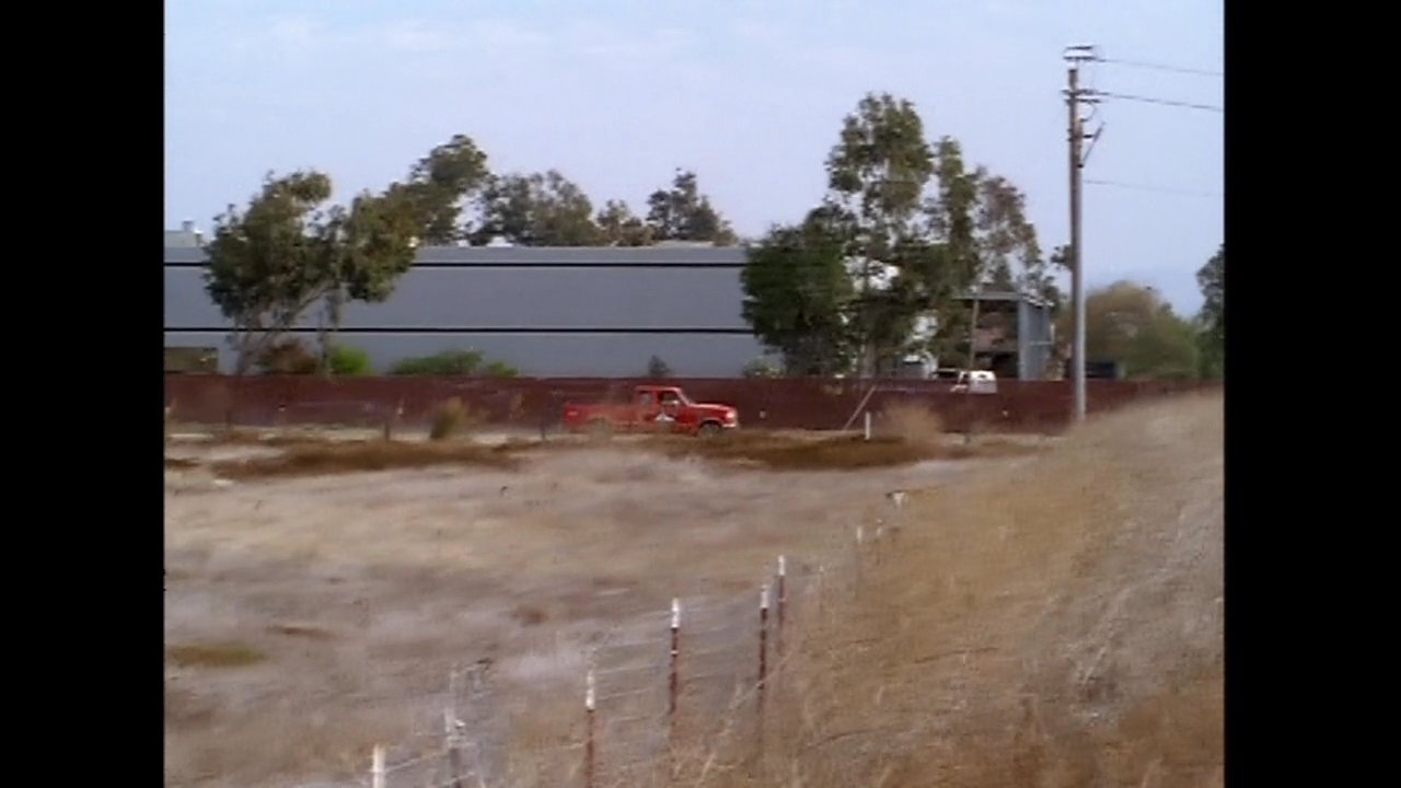 Individuals in an office environment observe a red truck driving along a dirt road near an industrial facility.
