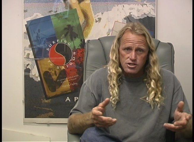 A long-haired professional surfer sits in a studio setting, animatedly discussing experiences and technical aspects of surfing through expressive hand gestures.