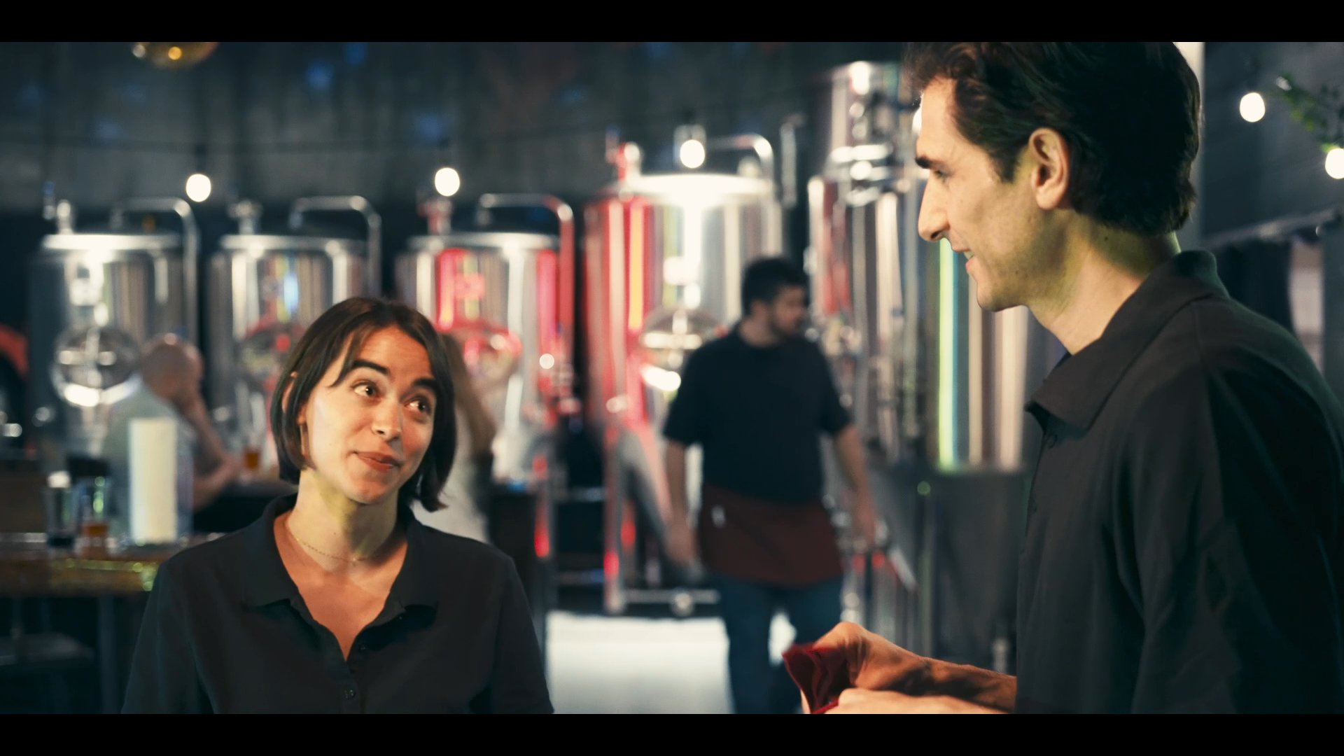 Two staff members engage in a professional yet friendly conversation within an industrial-style brewery environment. The setting features prominent stainless steel fermentation tanks in the background, underscoring the operational context.