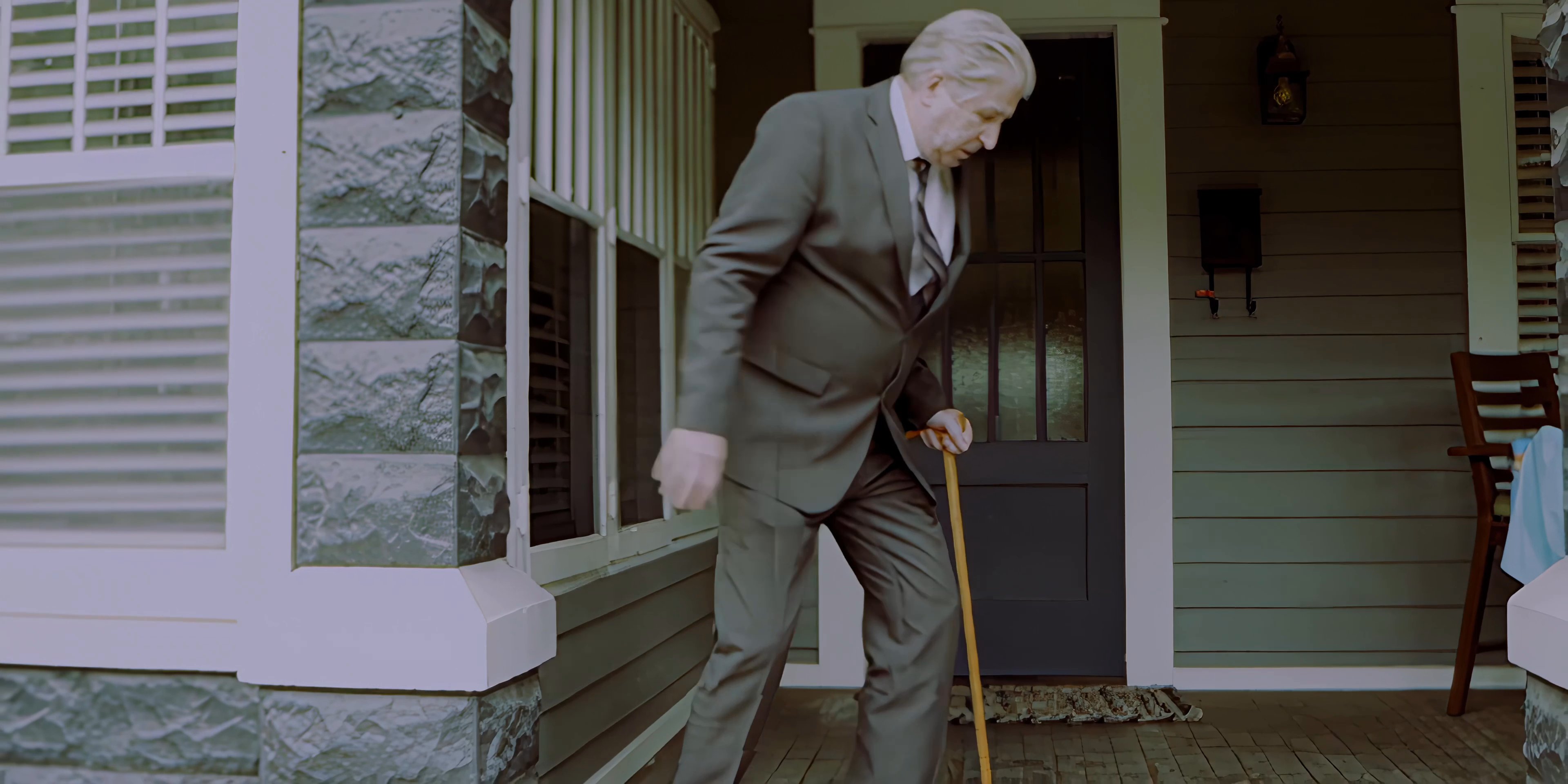 An elderly man in a suit uses a cane to step out of his house onto the porch. A younger man waits nearby outside, observing the transition.