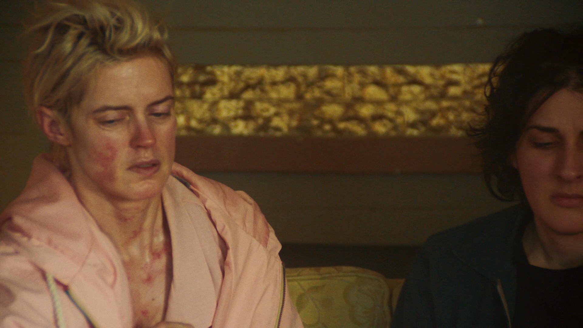 Two individuals sit on a sofa in a dimly lit room, appearing distressed and weary while a man observes them. The scene captures a moment of quiet conflict and palpable emotional strain within a residential environment.