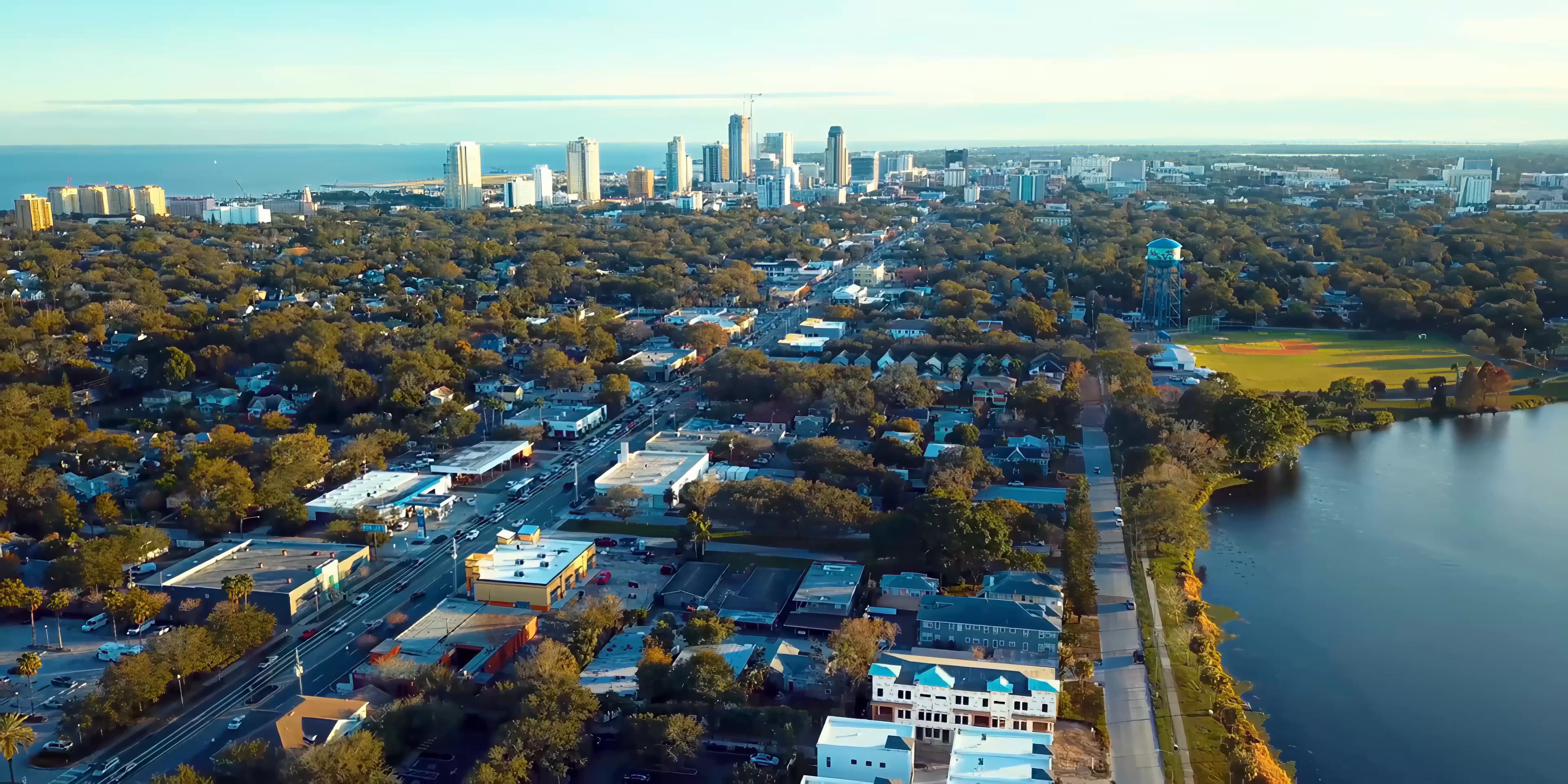 Two men engage in a professional dialogue while riding in the backseat of a vehicle. The scene transitions to a sweeping aerial perspective of a coastal city and a suburban street corner.