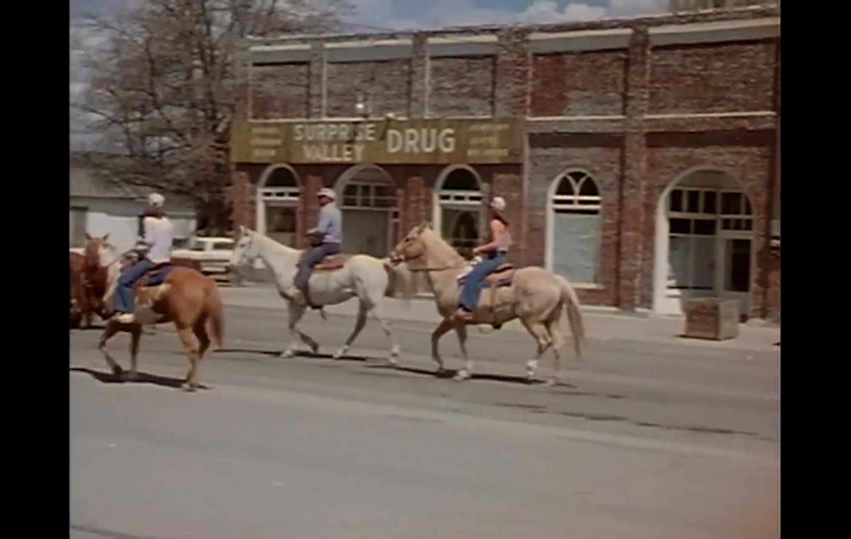 Ranchers and horseback riders guide a herd of cattle along a small-town main street, passing storefronts and a parked pickup. Snow-capped mountains rise behind the town as the herd moves through the intersection.