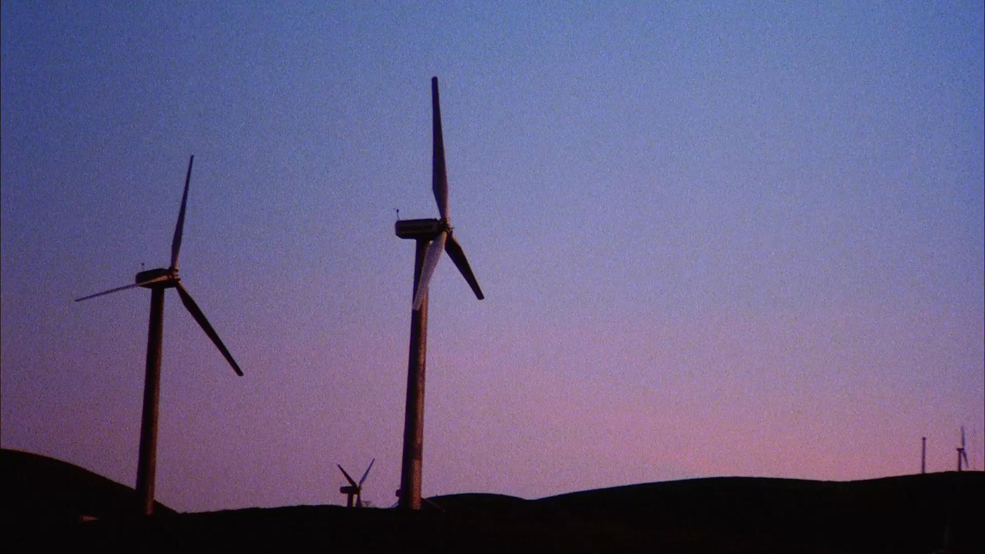 Fragmented aircraft debris lies on an asphalt road near a vast wind farm. A hiker with a backpack and beanie observes the landscape at dusk.