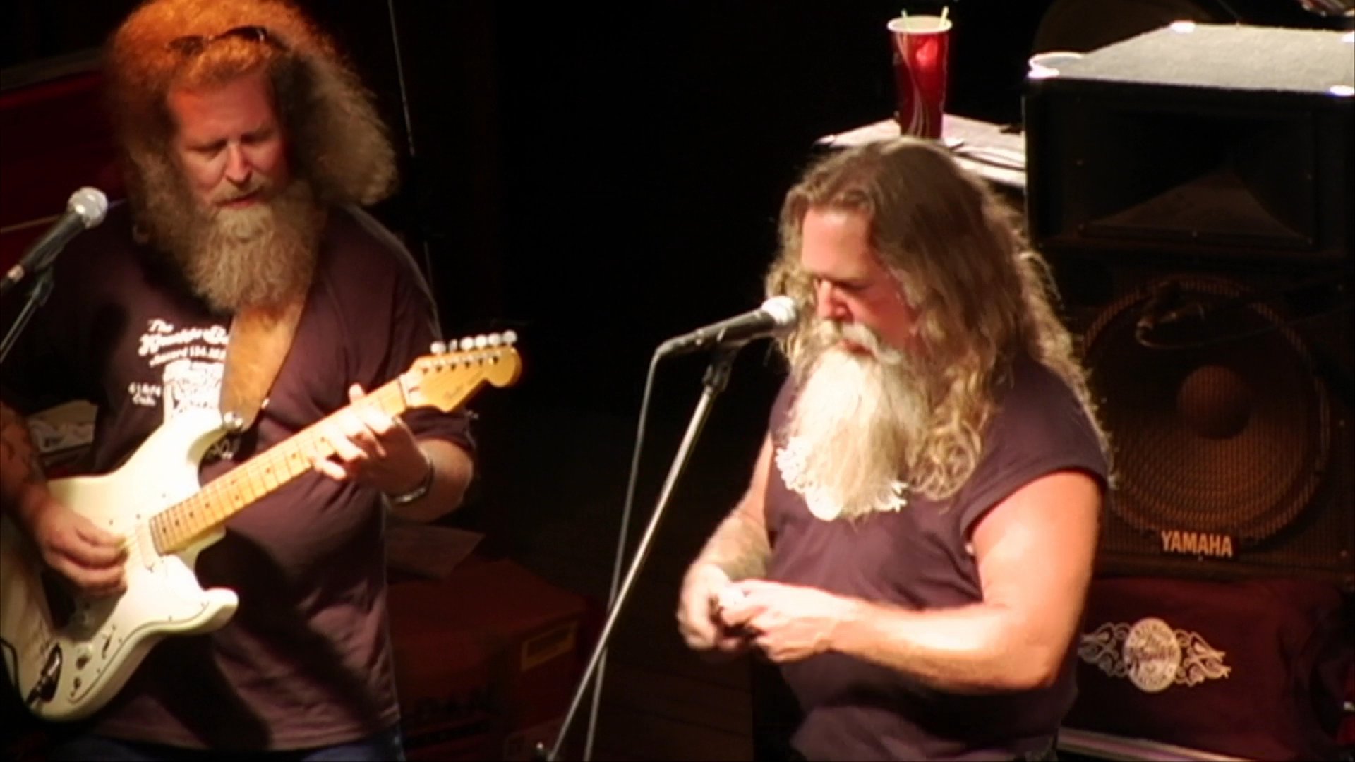 A man with long hair and a beard plays an electric guitar and harmonica during a live performance, while a separate segment features a brief interaction between two individuals.