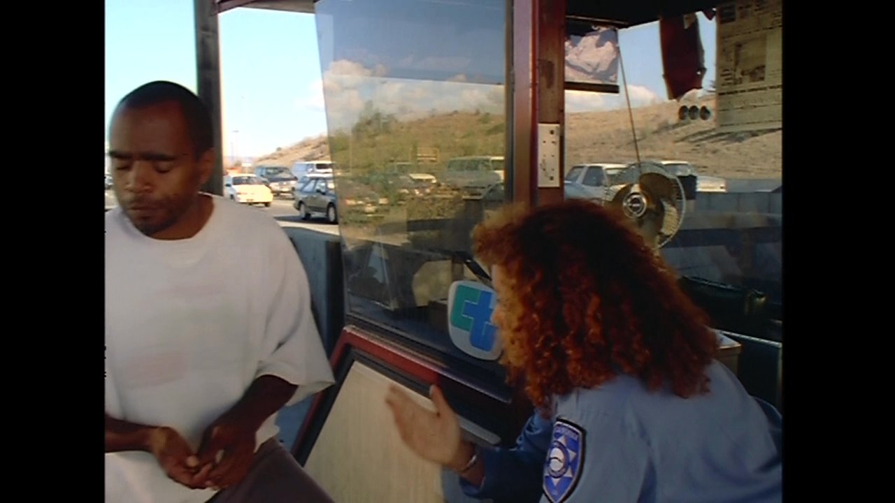 A man hands cash to a toll booth attendant through a service window. Following the exchange, he walks away from the booth.