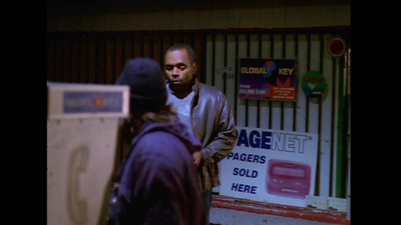 A man in a leather jacket initiates a phone call at an outdoor payphone kiosk during the night. He stands on a city sidewalk while another individual waits nearby.