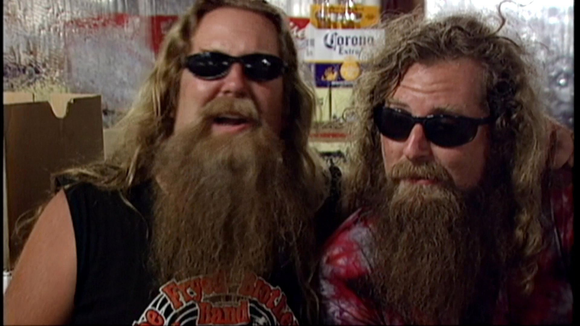 Two long-haired men with full beards and sunglasses engage in conversation and share a lighthearted, jovial moment during an interview.