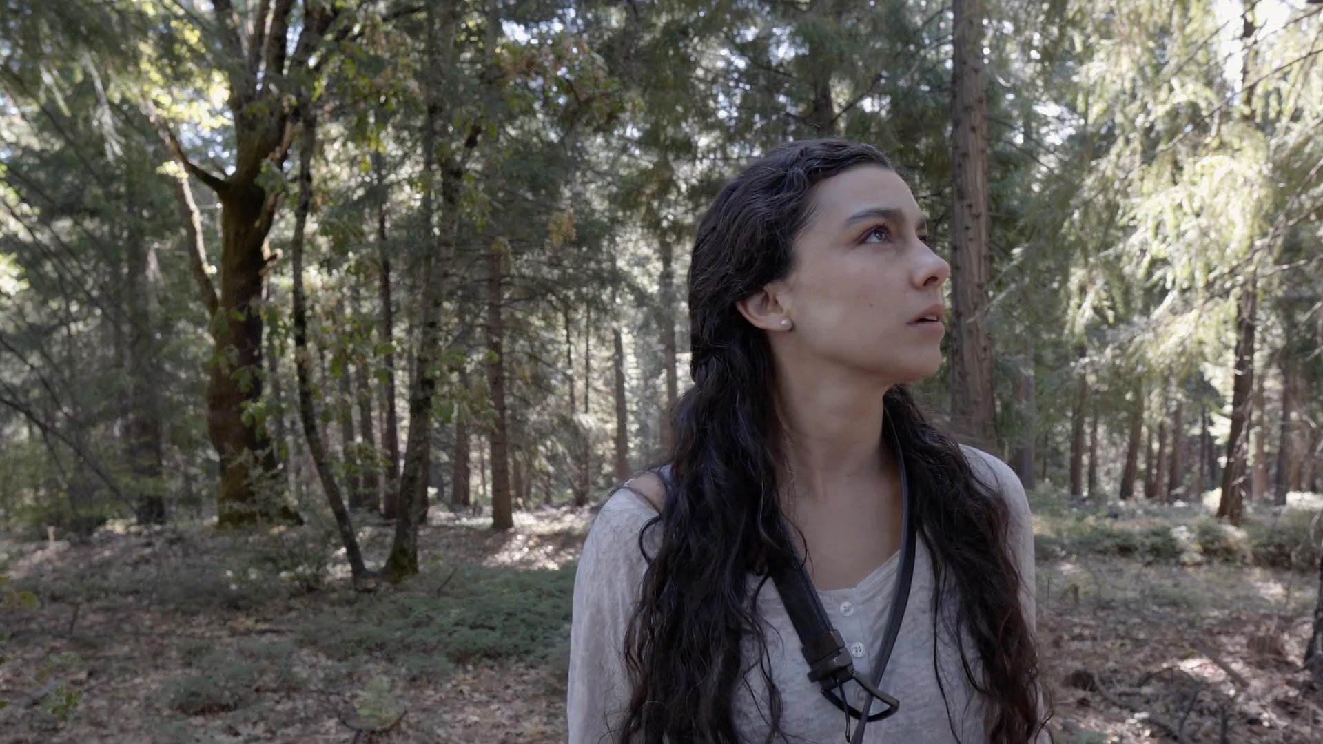 A woman cautiously scans her surroundings in a wooded area before discovering a small drone hovering nearby. Her expression transitions from inquisitive concern to focused observation as she tracks the aerial device.