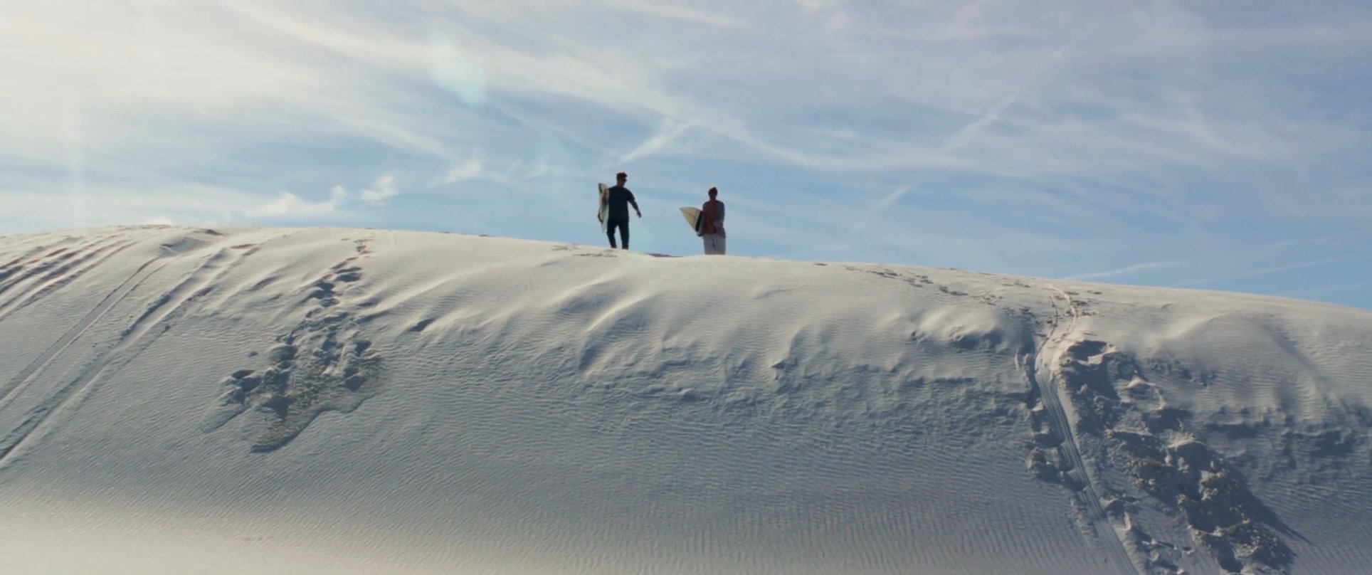 Two individuals carry surfboards while traversing a vast, sunlit white sand desert landscape. The pair walks along the dunes before stopping to take in the panoramic view.