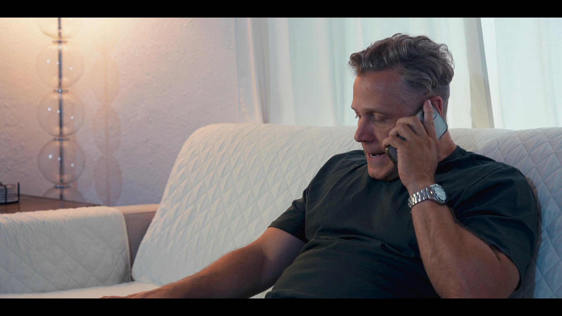 A man engages in a focused phone call while relaxing on a sofa, transitioning to aerial footage of a vehicle navigating a suburban residential road. The sequence juxtaposes personal communication with a daily travel routine.
