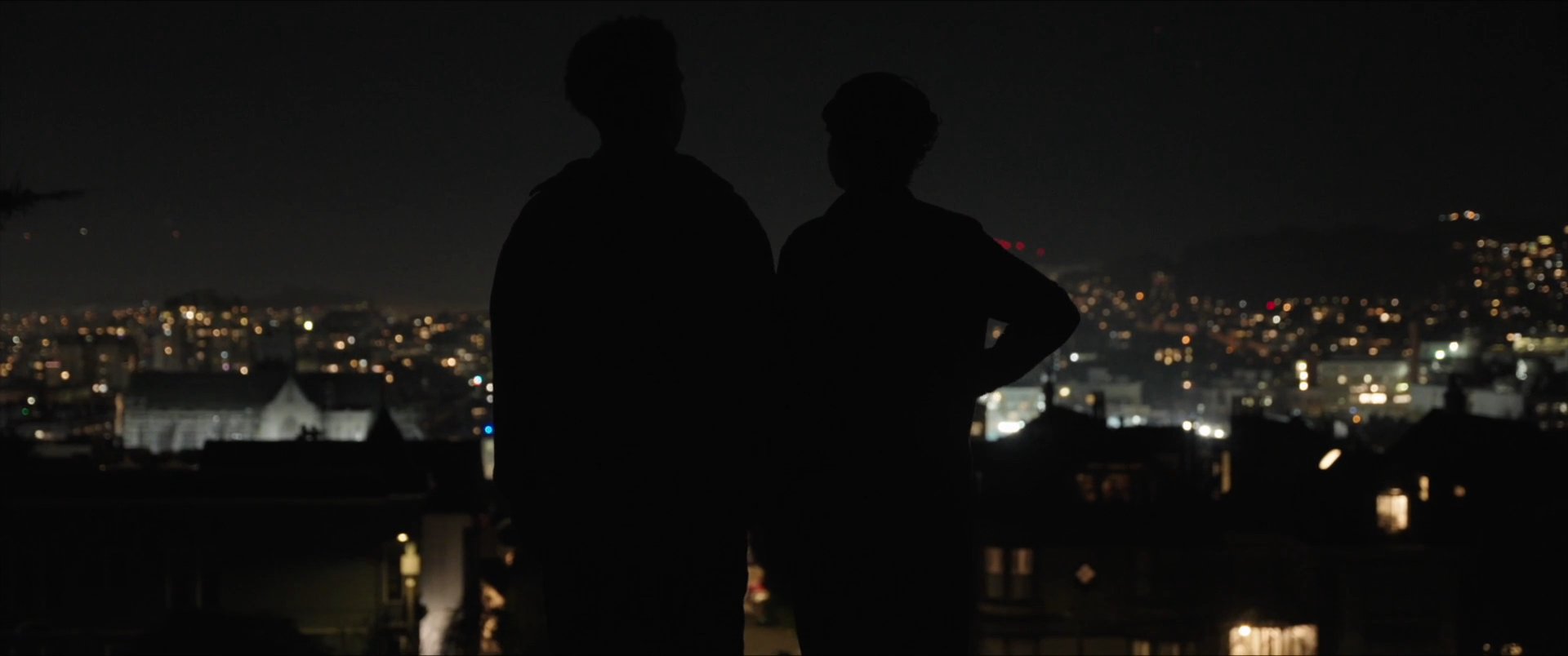 Two individuals stand on a vantage point overlooking a city at night while engaged in conversation. The scene transitions between close-up shots of the pair and wide panoramic views of the urban landscape illuminated by a full moon.