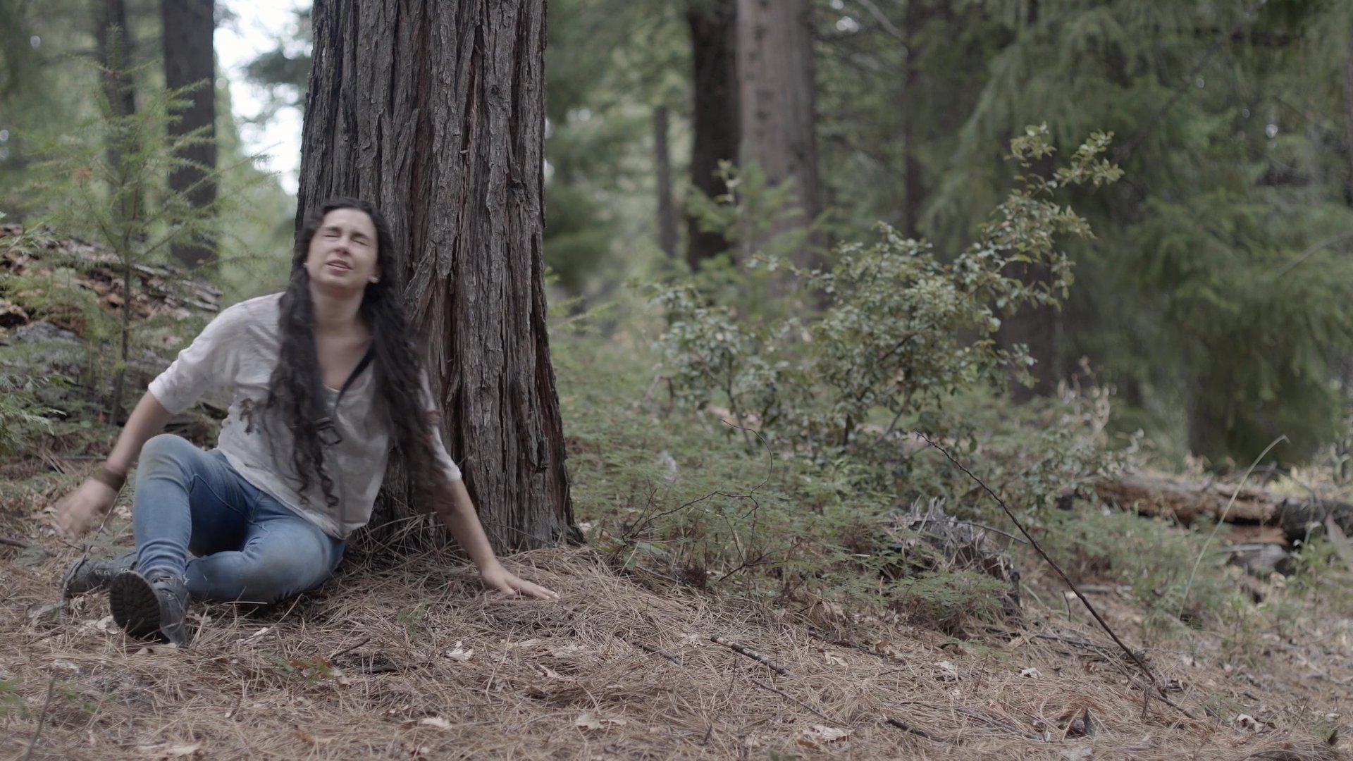A woman crawls across the forest floor before resting against a large tree, showing signs of exhaustion and eventual calm. The sequence captures a moment of physical struggle transitioning into a state of reflection in a wooded environment.