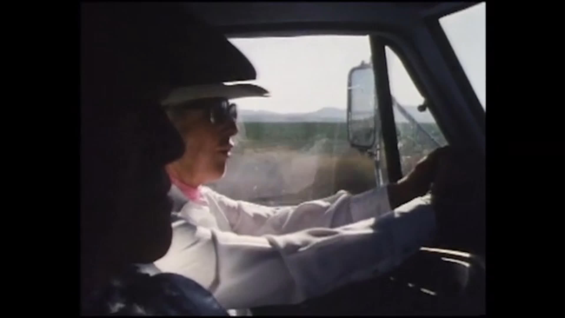 An older woman drives through open countryside, speaking warmly about enjoying her work and the people she has worked alongside. She reflects that wealth was never her goal as the rural landscape passes outside the truck.