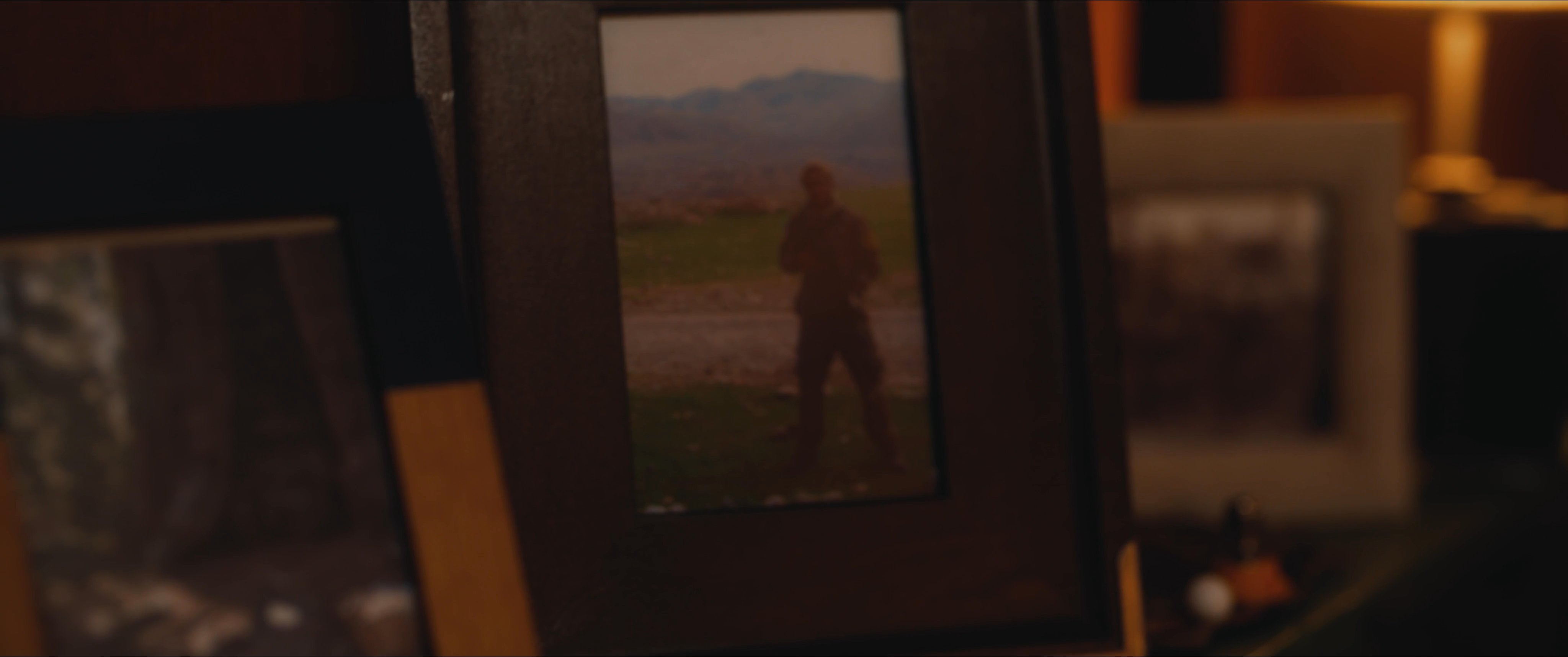 A man wanders through a dark, wooded area toward a secluded cabin before retreating inside to reflect on framed personal photographs amidst an array of empty bottles.
