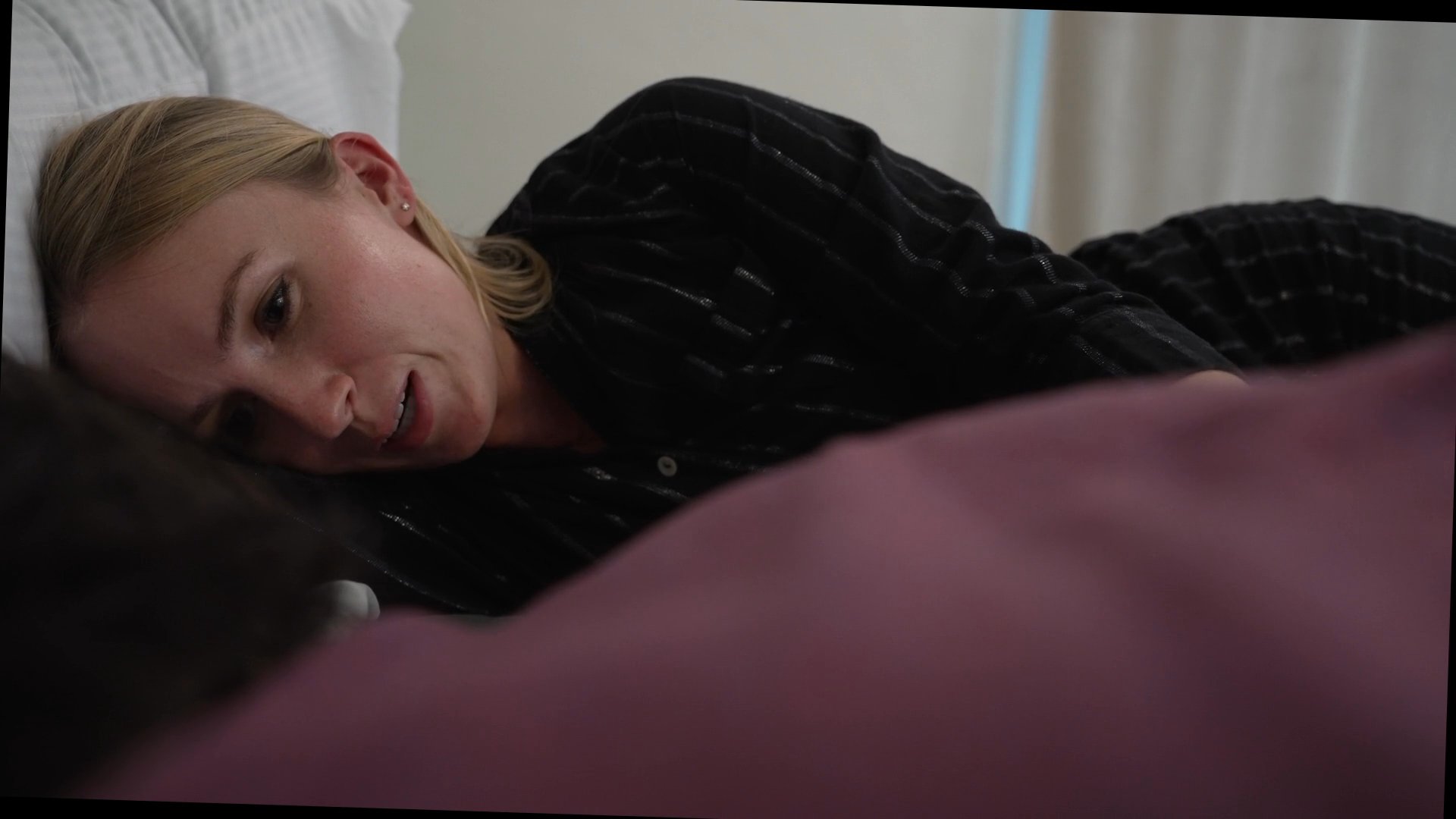 A woman rests comfortably in bed and interacts affectionately with a partner out of view before transitioning to him lying in the same bed.