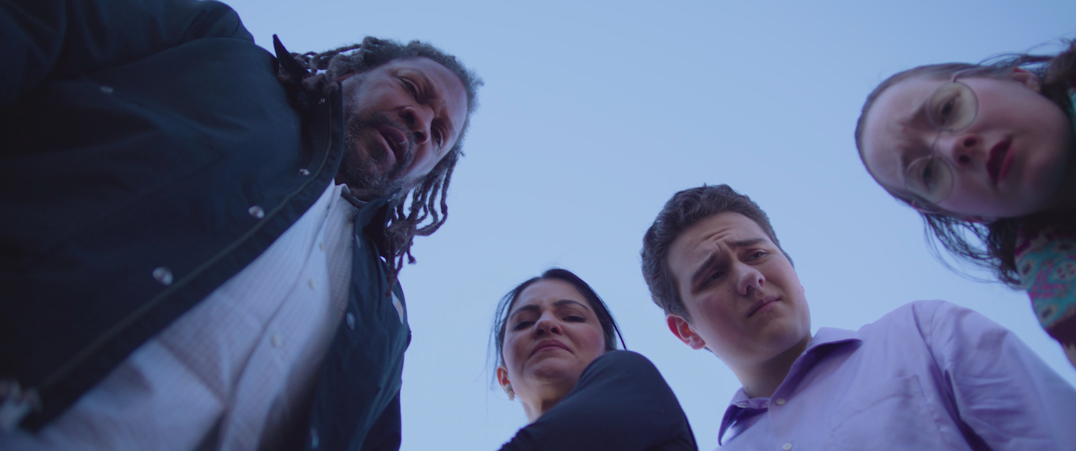 A group of people reacts with shock and dismay after uncovering a human hand buried in a dirt yard. They stare down at the object, visibly disturbed by the sudden, grim discovery.