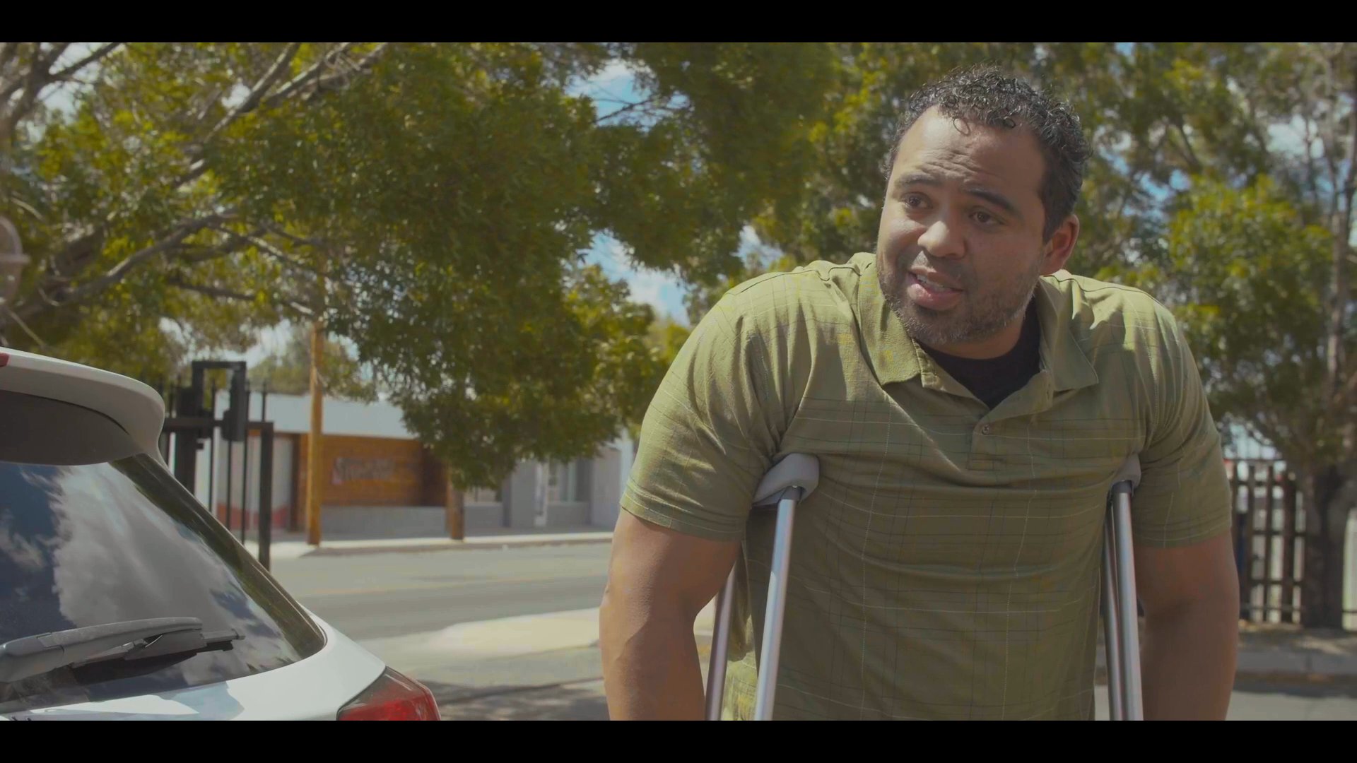 A woman in a maroon shirt and a man using crutches engage in a serious, focused conversation beside a parked vehicle in an outdoor parking lot.
