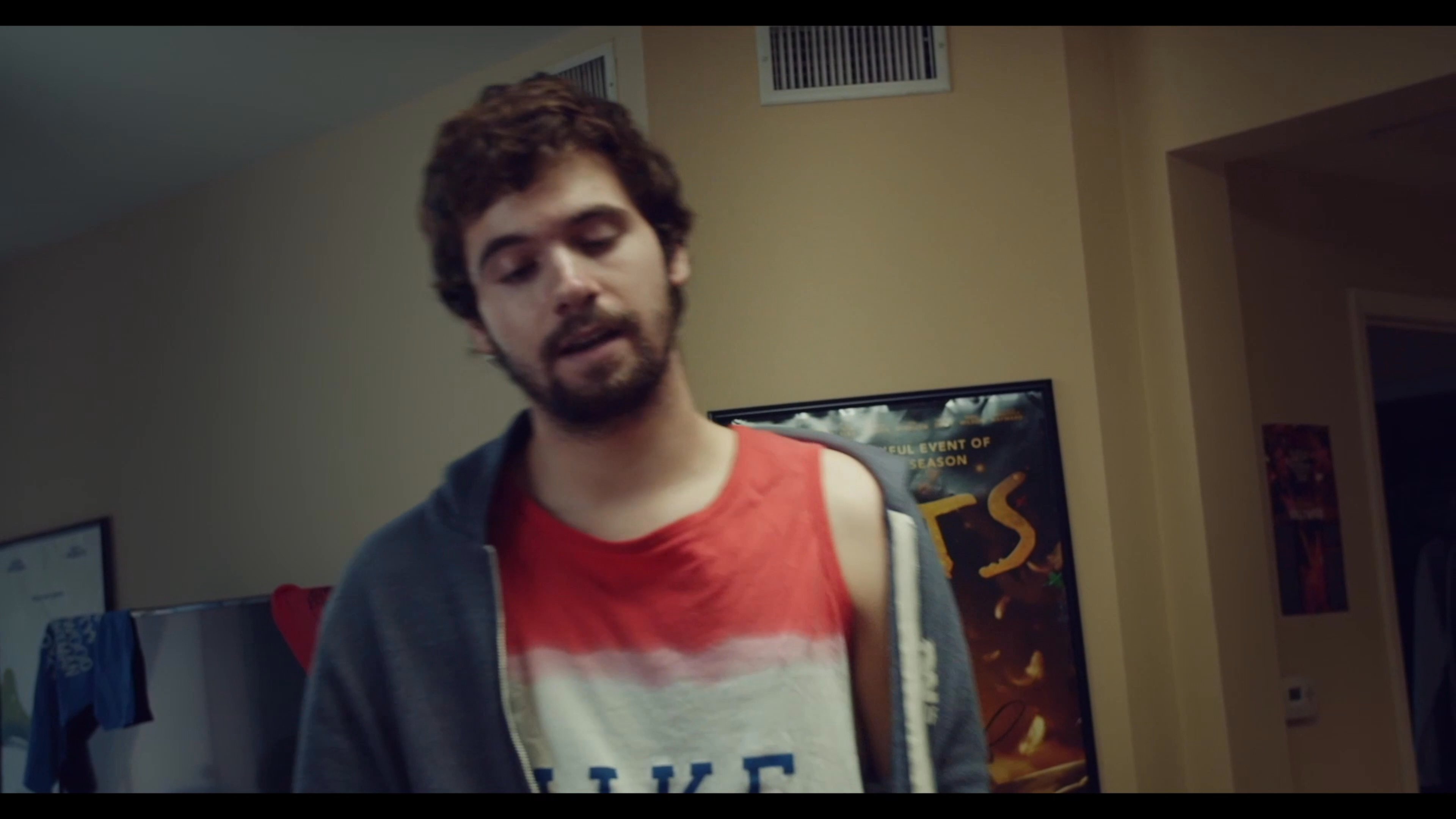 A man in a patterned shirt reacts with shock and frustration while another man sits on the floor crying amidst a cluttered apartment. The scene depicts a tense emotional confrontation between two individuals.